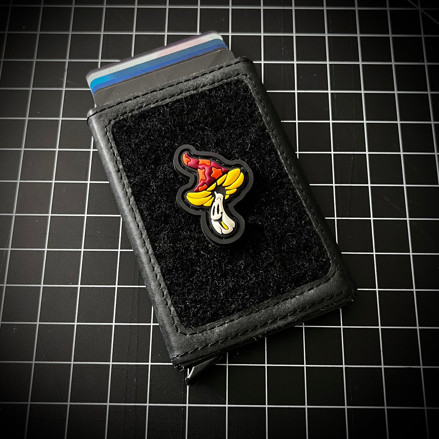 Shroomie RE - Fire PVC ranger eye morale patch of a smiling mushroom character with an orange and yellow cap on a velcro wallet with a grid background.