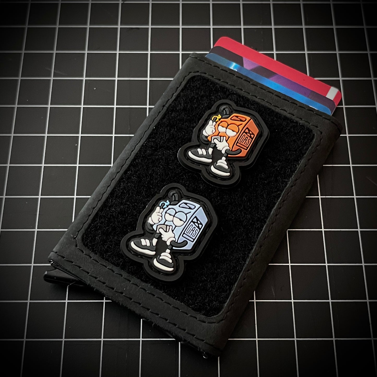 Fueled Up RE - Red PVC ranger eye morale patch of a red cartoon gas cannister character holding up an orange lit match in one hand and a lighter in the other on a velcro wallet and grid background.