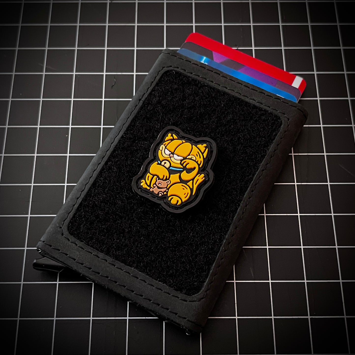 Garfield Neko RE PVC ranger eye morale patch of an orange cat holding a teddy bear sitting in a lucky cat maneki-neko pose on a velcro wallet with a grid background.