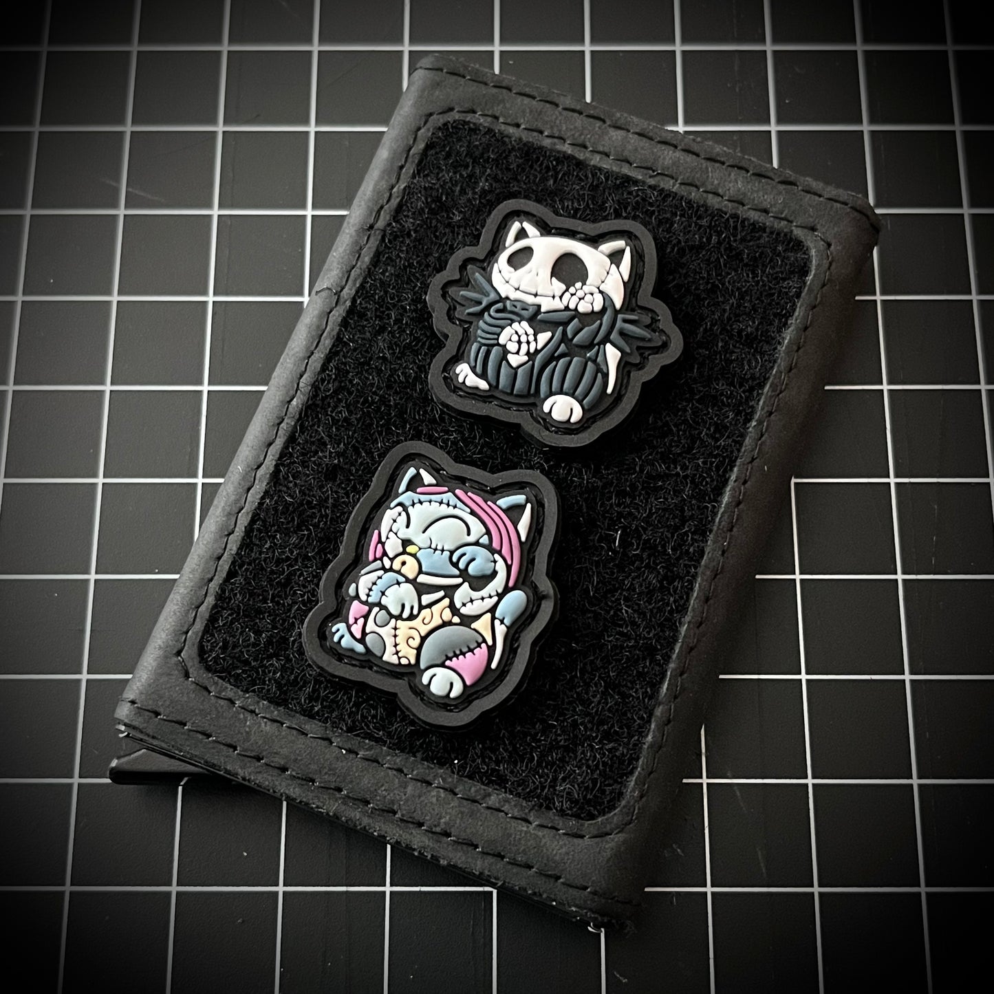 Jack & Sally Neko RE Set PVC ranger eye morale patches of a skeleton character in a black suit and blue patchy girl sitting in a lucky cat maneki-neko pose on a velcro wallet and a grid background.