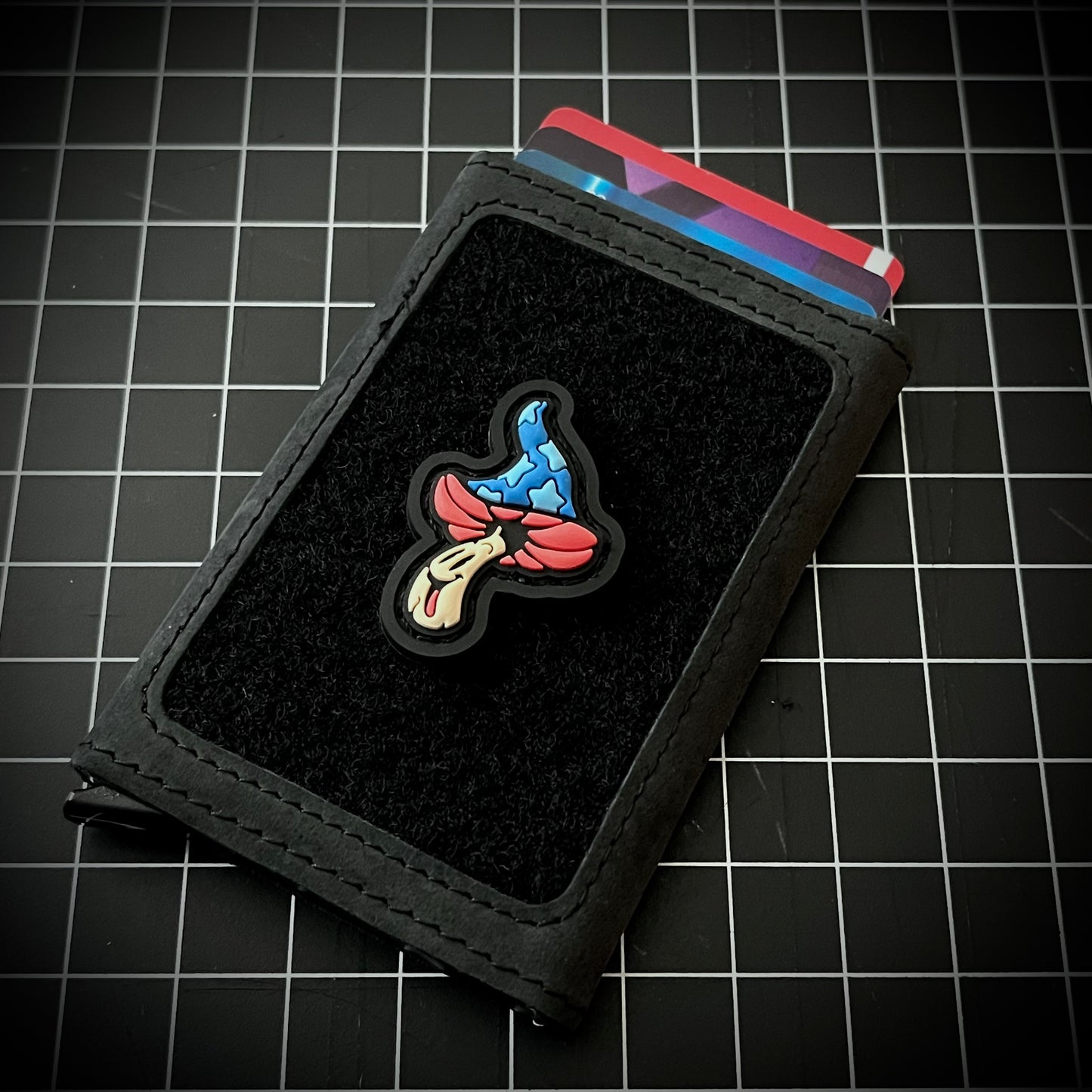 Shroomie RE - Fantasia PVC ranger eye morale patch of a red, blue, and white smiling mushroom whose cap is reminiscent of mickey sorcerer fantasia on a velcro wallet and a grid background