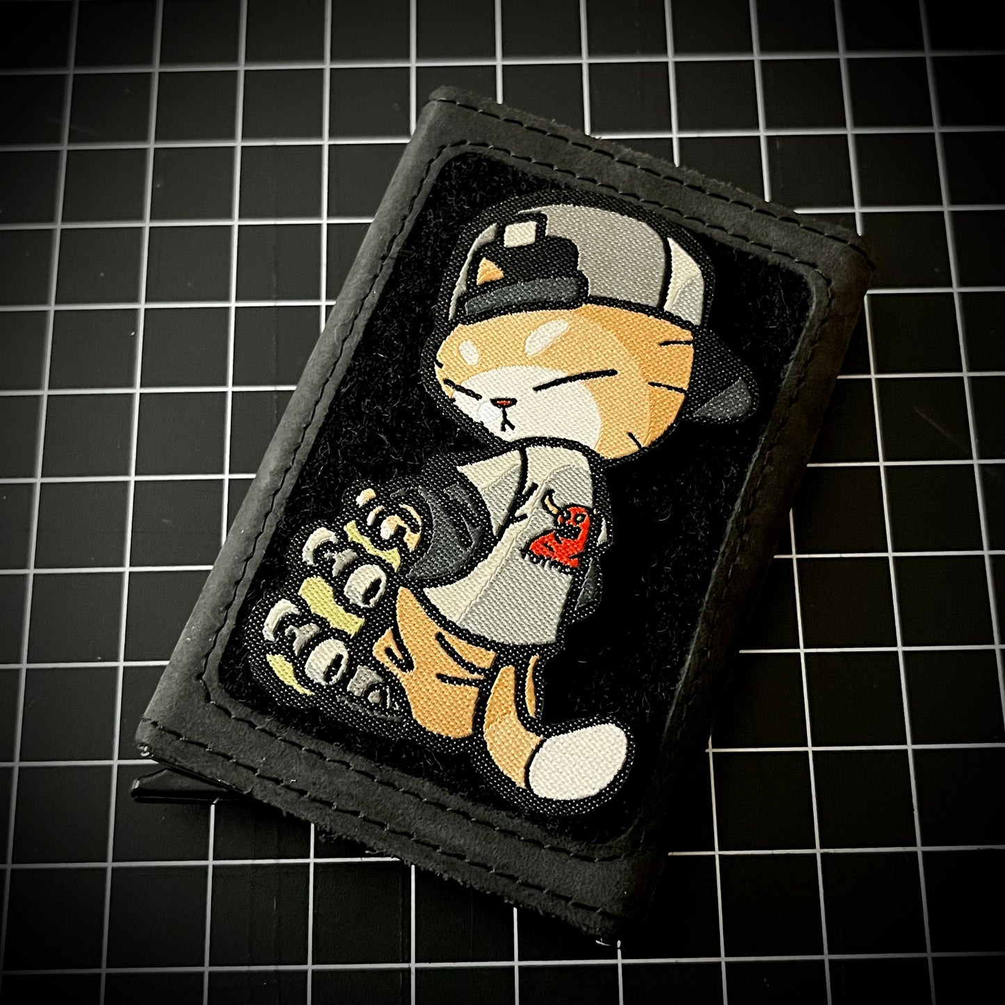Skate Cato Patch woven nylon morale patch of an orange cat wearing a backwards hat and Toy Machine shirt holding a skateboard on a wallet and a grid background.
