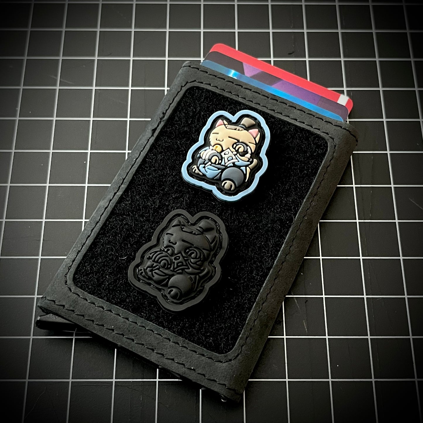 Sokka Neko RE and Sokka Neko Blackout RE PVC ranger eye patches of a cartoon character on a velcro wallet with a grid background.