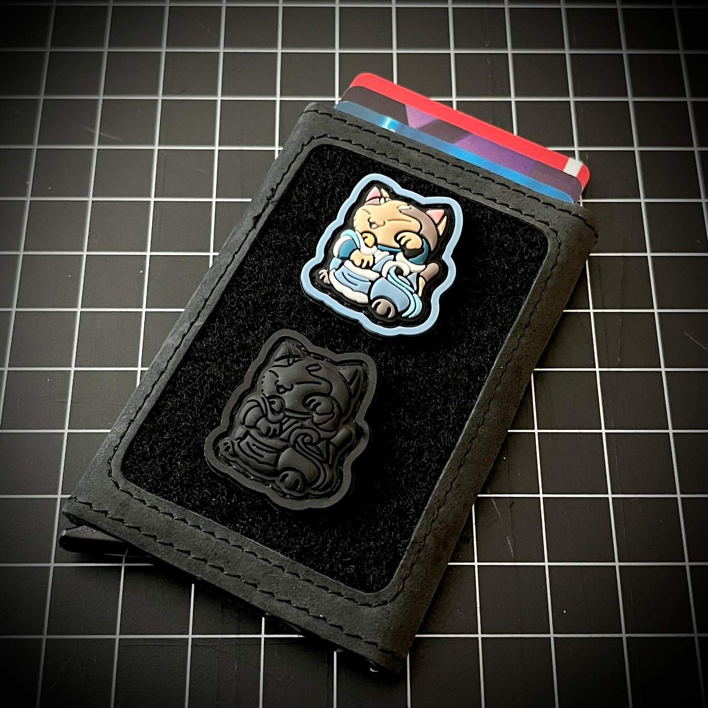 Water Bender Neko RE and neko Blackout RE of a cartoon character sitting in a lucky cat maneki-neko pose on a velcro wallet with a grid background.