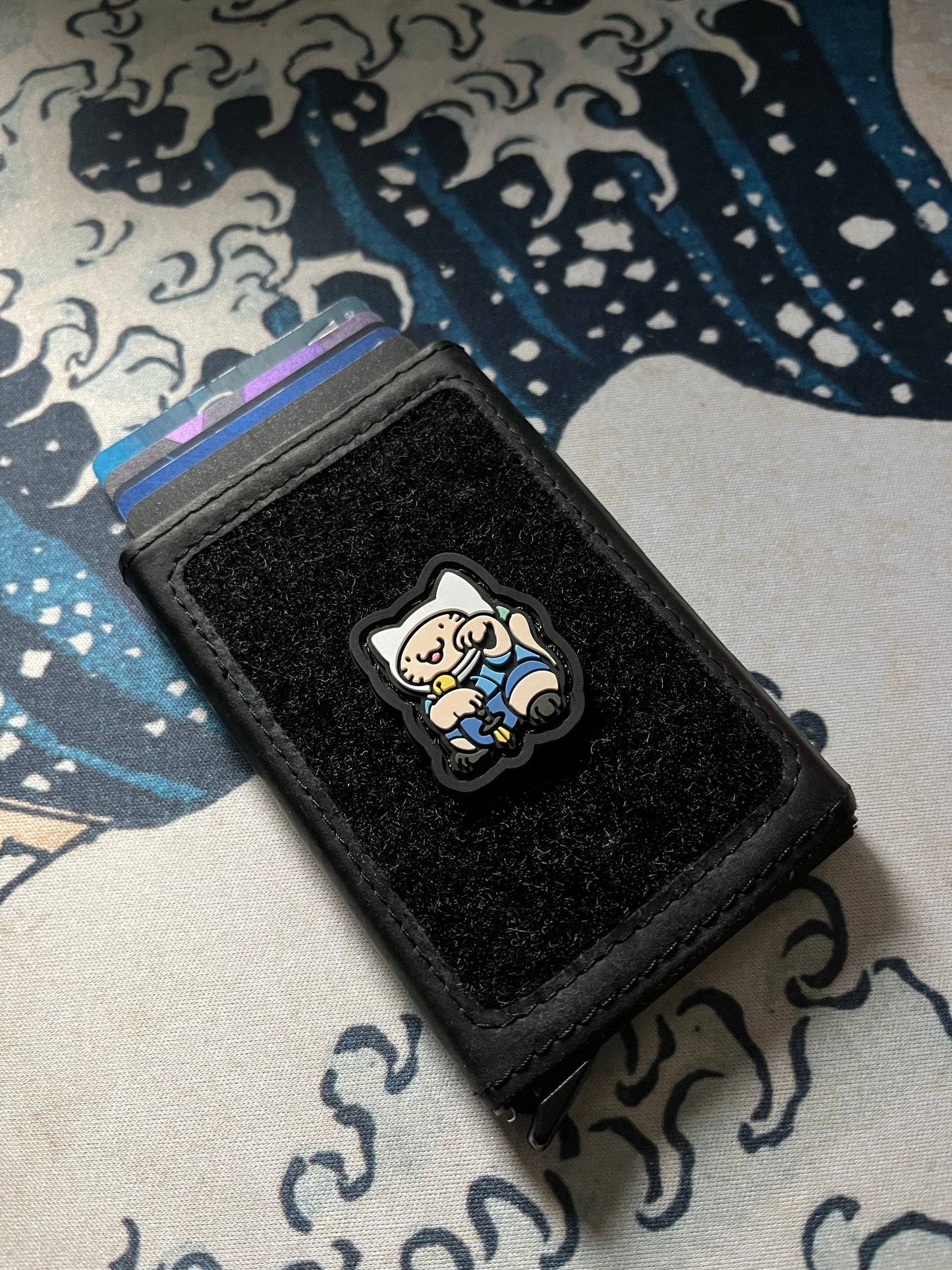 Adventure Neko RE ranger eye patch with a cartoon cat character wearing a white hat and blue outfit in a lucky cat maneki neko pose on a black wallet with a Great Wave background.