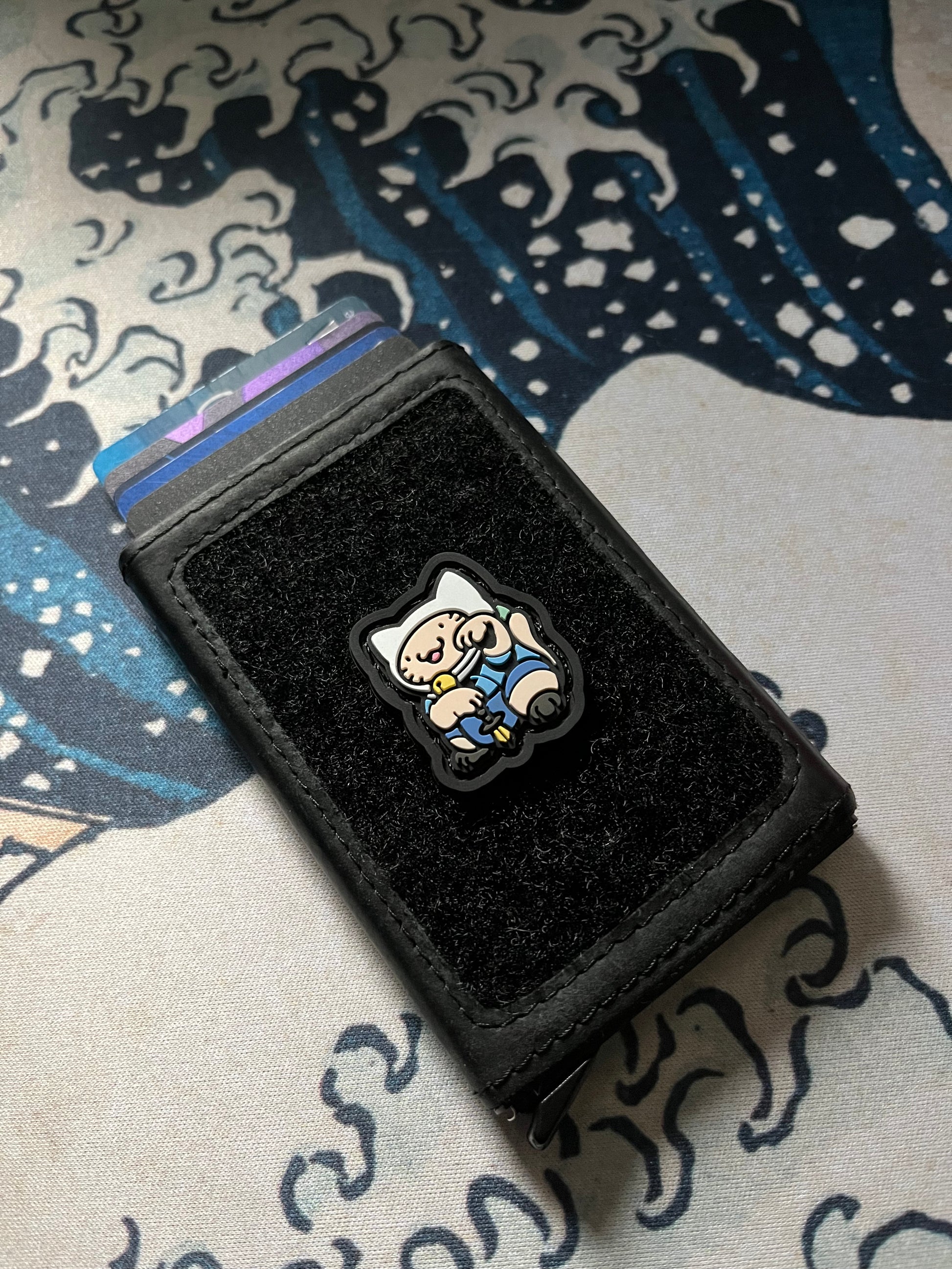 Adventure Neko RE ranger eye patch with a cartoon cat character wearing a white hat and blue outfit in a lucky cat maneki neko pose on a black wallet with a Great Wave background.