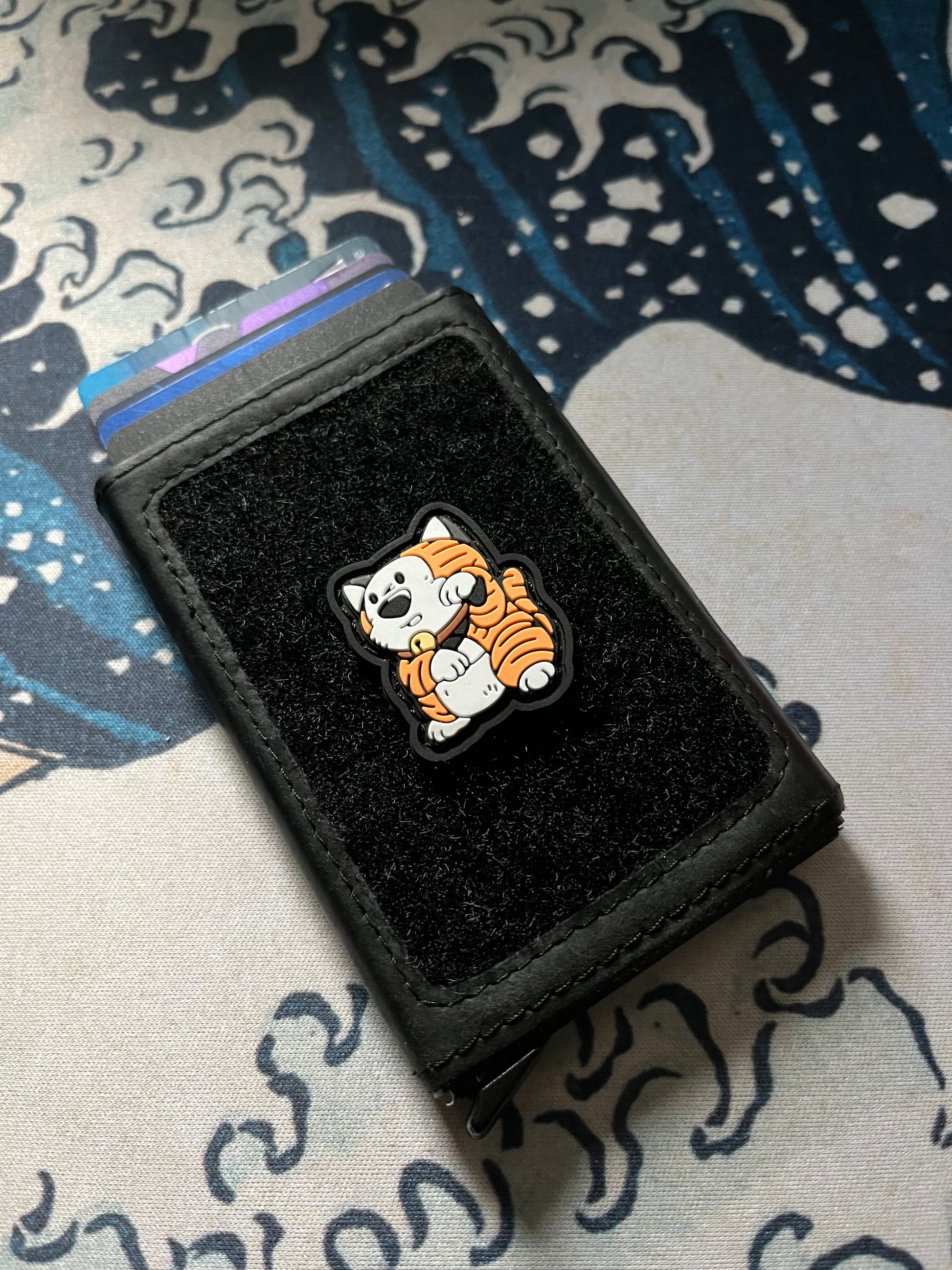 Comic Tiger Neko RE ranger eye patch depicting a cartoon tiger in a lucky cat maneki neko pose on a velcro wallet with a Great Wave background.