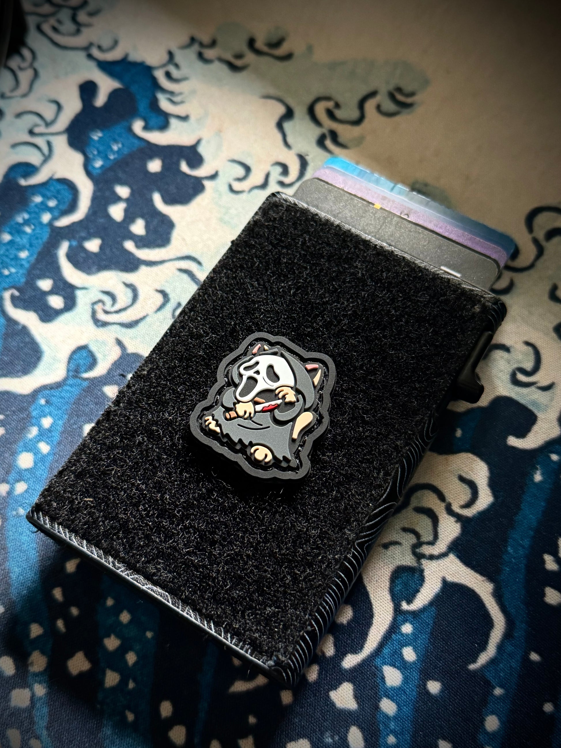 Ghost face Neko RE Halloween patch depicting a cat wearing a black dobe, white mask, holding up a knife in a maneki nekolucky cat pose on a wallet with a Great Wave painting background.