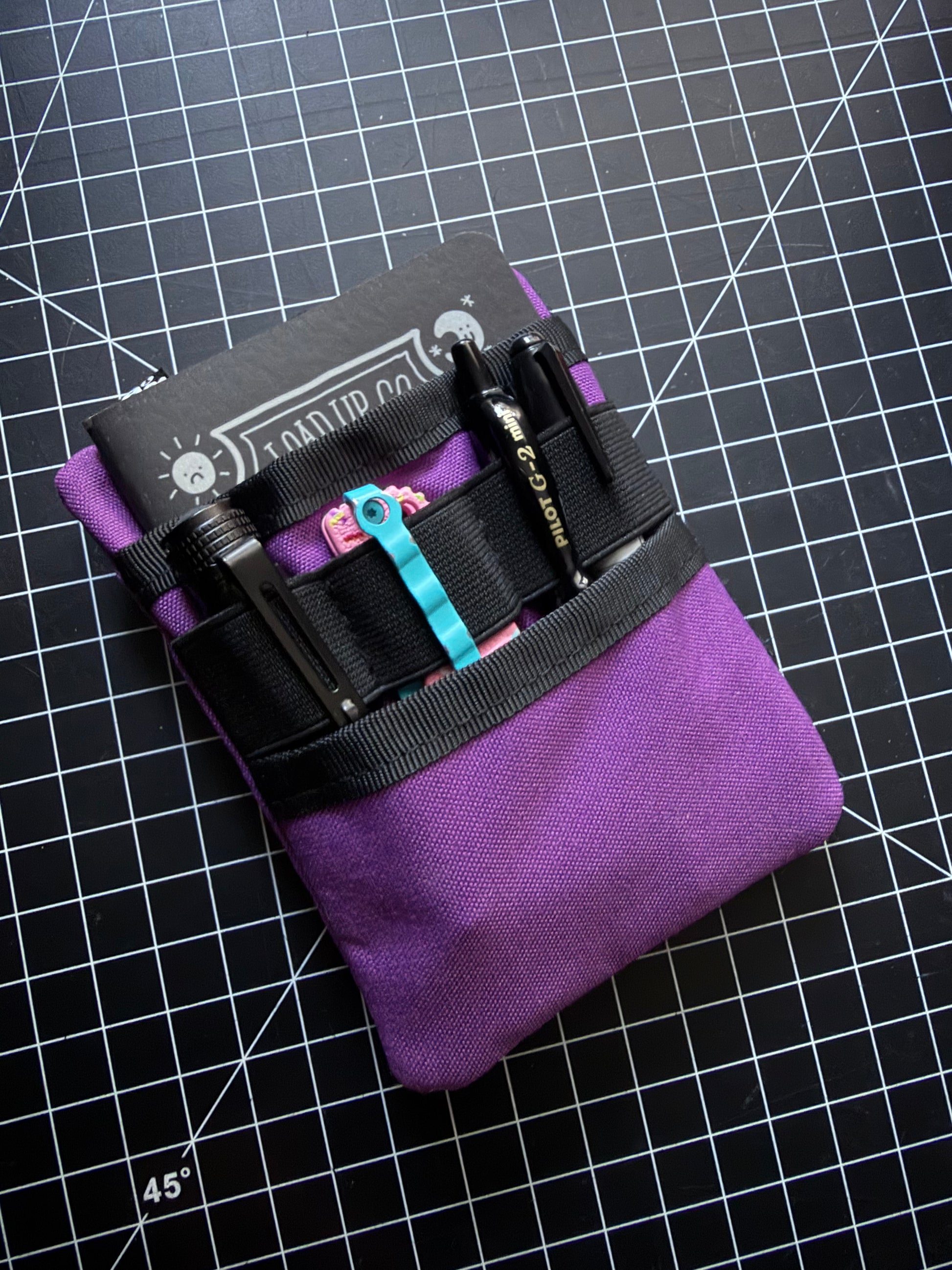 A purple canvas utility pouch with black elastic bands holding pens and tools, photographed on a black gridded cutting mat.