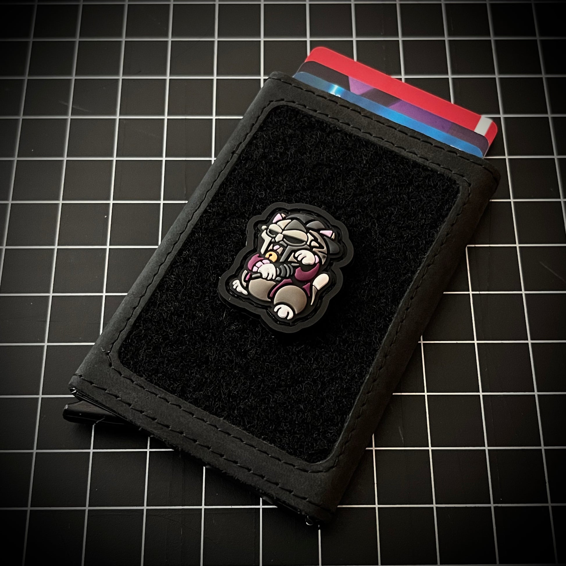 MF Doom Neko RE PVC ranger eye morale patch of a rapper sitting in a lucky cat maneki-neko pose on a velcro wallet and grid background.