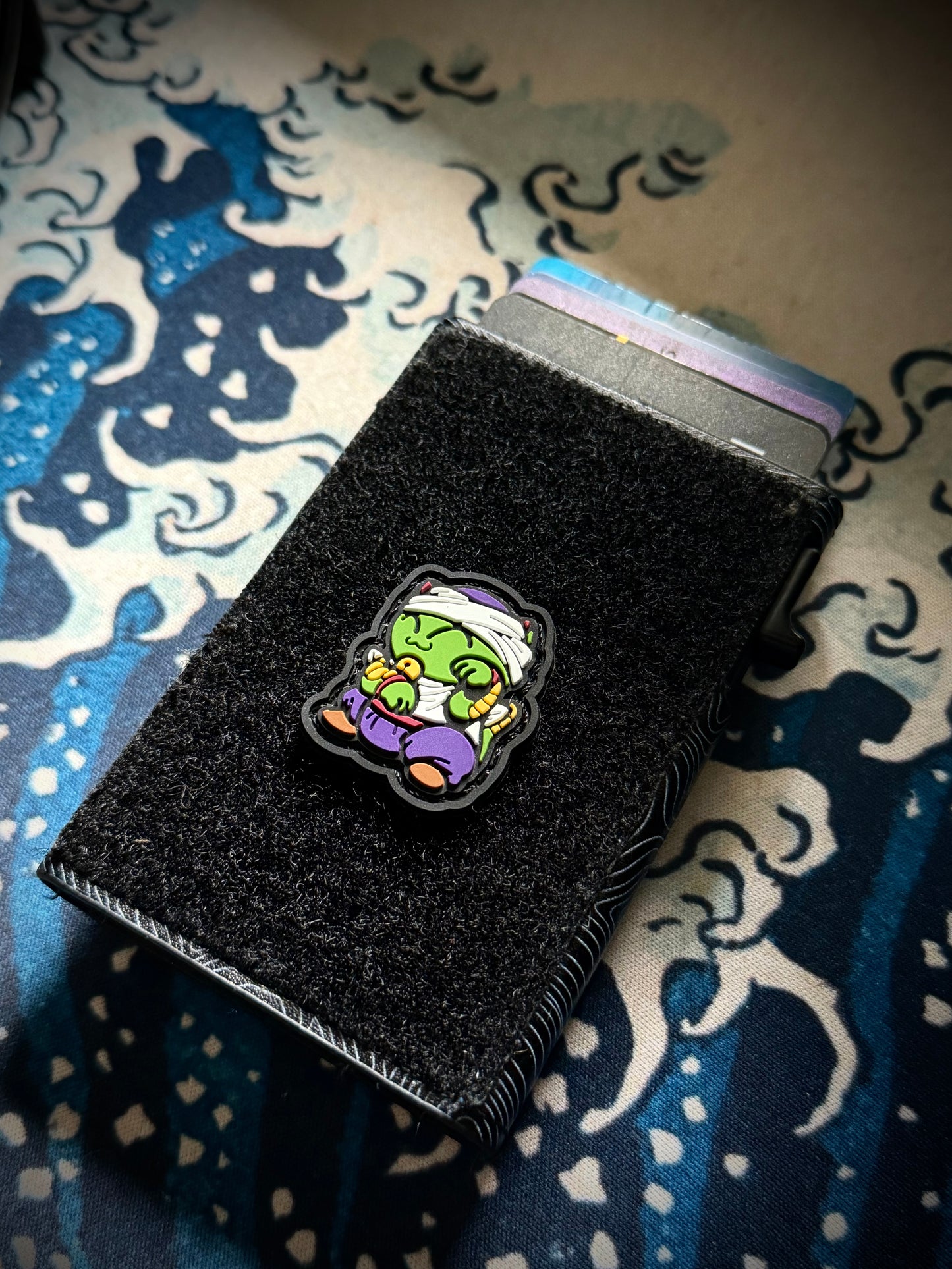 Piccolo Neko RE Colorful cartoon character with green skin, purple outfit, and white headband on a black border on a velcro wallet with a Great Wave background.