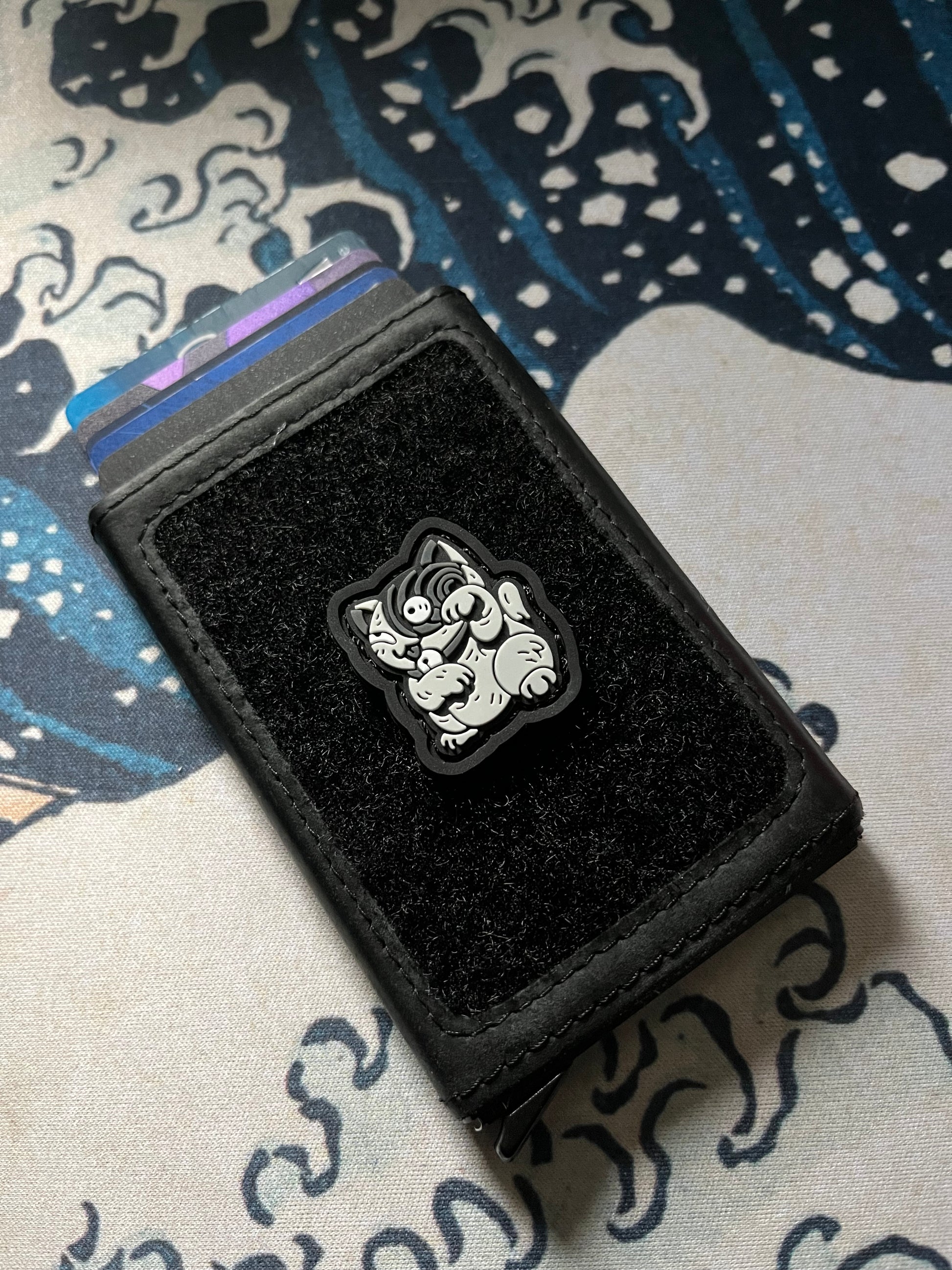 Uzumaki Neko RE ranger eye patch depicting a gray cat with a swirl on its face in a lucky cat maneko pose on a velcro wallet with a Great Wave background.