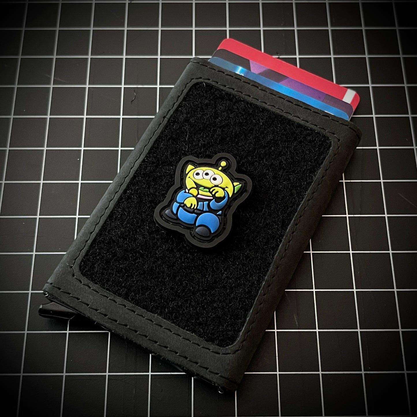 Alien Neko RE PVC ranger eye patch of a green alien with 3 eyes and a blue outfit sitting in a lucky cat maneki-neko pose on a velcro wallet and a grid background.