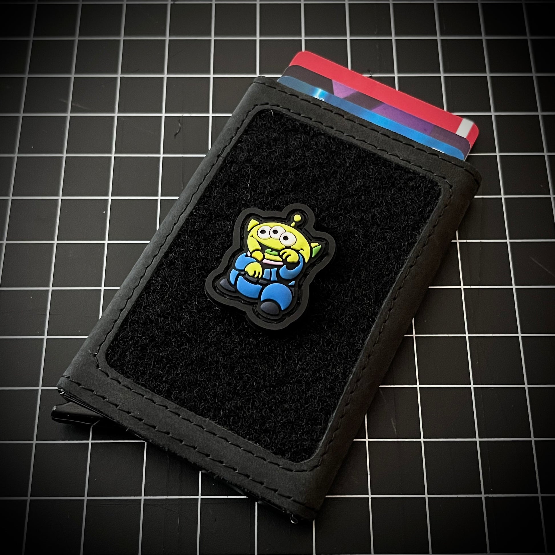Alien Neko RE PVC ranger eye patch of a green alien with 3 eyes and a blue outfit sitting in a lucky cat maneki-neko pose on a velcro wallet and a grid background.