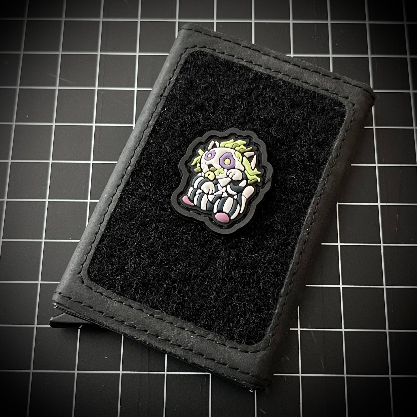 Beetlejuice Neko RE PVC ranger eye morale patch of a character with green hair and striped shirt sitting in a lucky cat maneki-neko pose on a velcro wallet and a grid background.