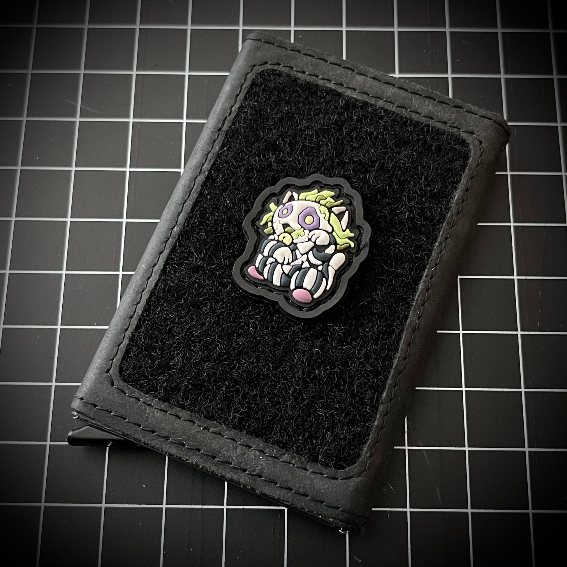 Beetlejuice Neko RE PVC ranger eye morale patch of a character with green hair and striped shirt sitting in a lucky cat maneki-neko pose on a velcro wallet and a grid background.