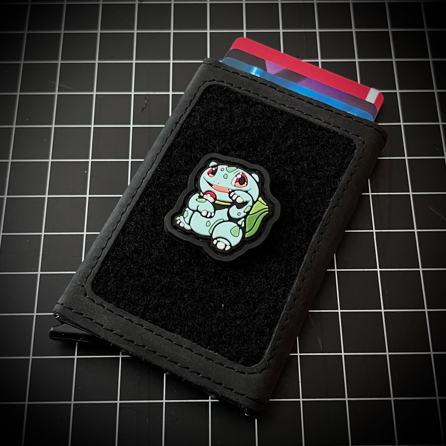 Bulbasaur Neko RE PVC ranger eye morale patch of a green character in a lucky cat maneki-neko pose on a velcro wallet and grid background.