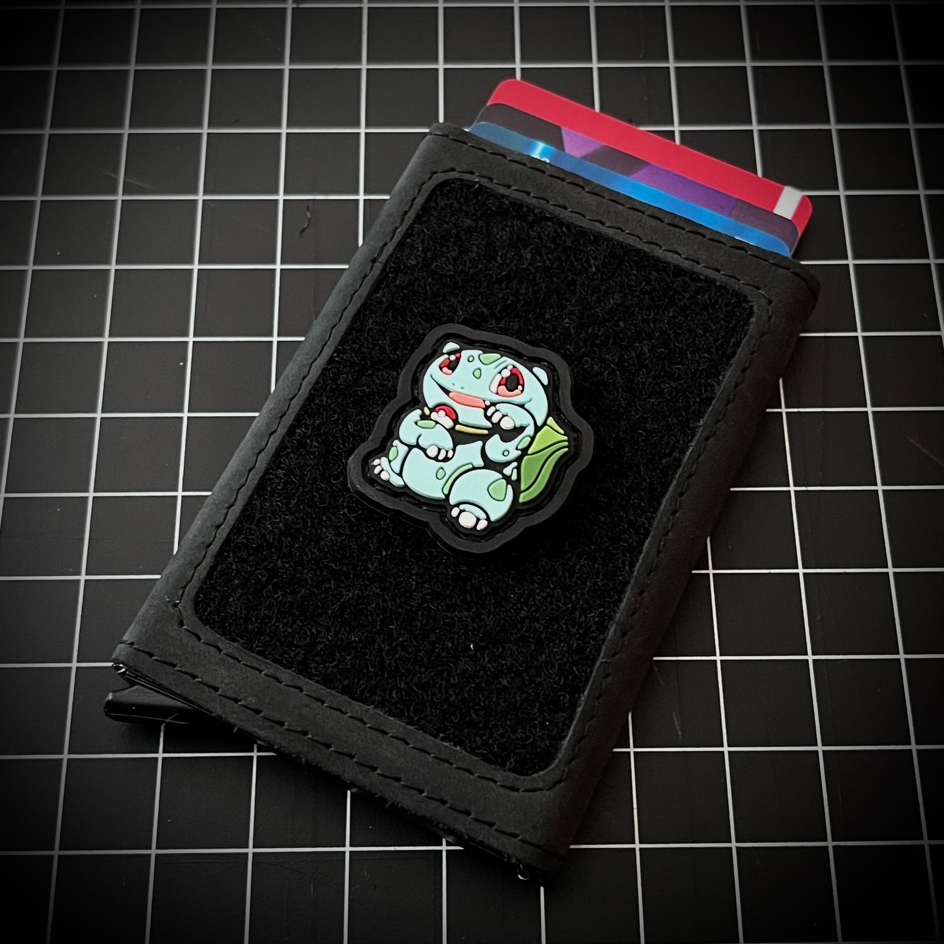 Bulbasaur Neko RE PVC ranger eye morale patch of a green character in a lucky cat maneki-neko pose on a velcro wallet and grid background.