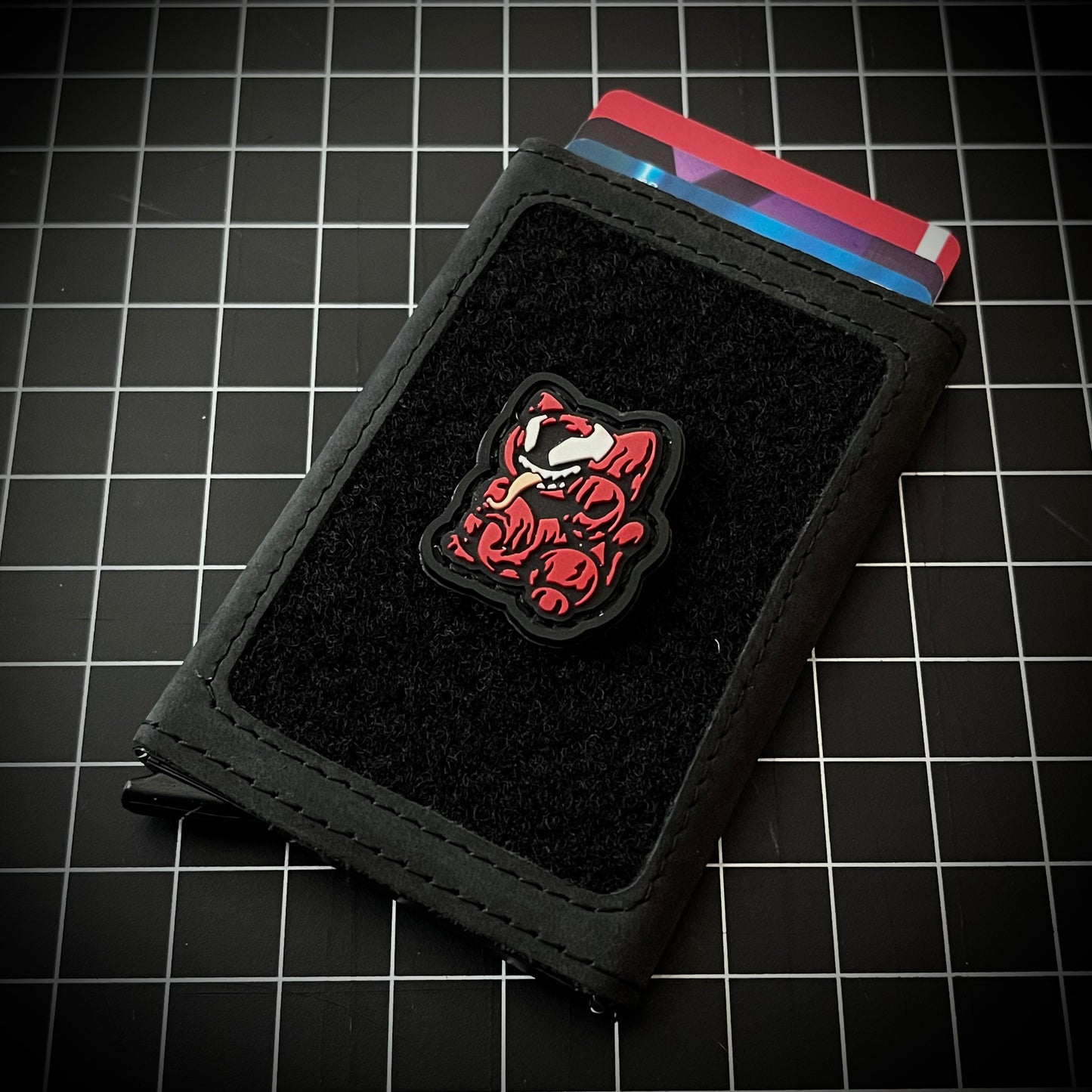Carnage Neko RE PVC ranger eye morale patch of a red cartoon character with its tongue sticking out sitting in a lucky cat maneki-neko pose on a velcro wallet and grid background.