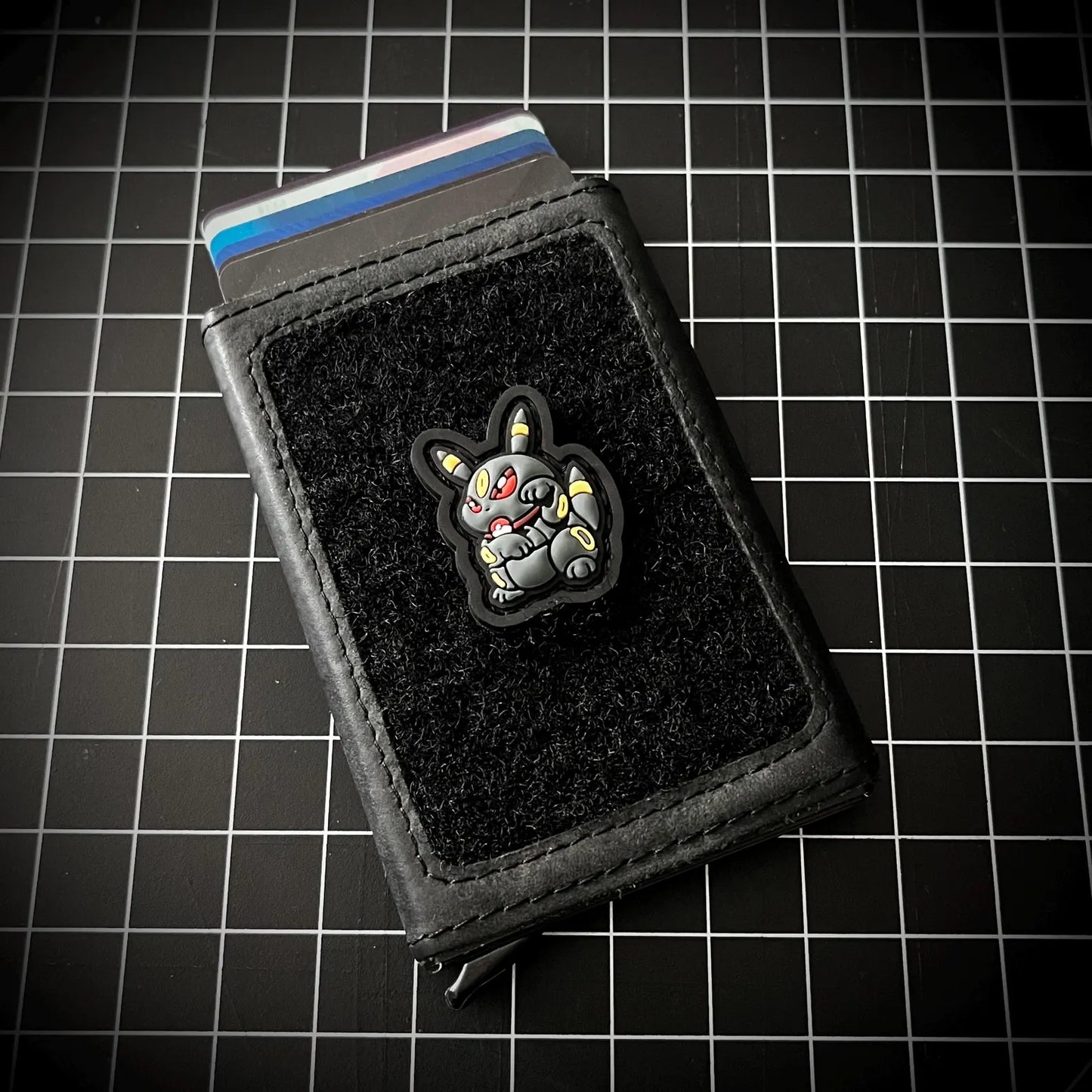 A black leather card holder wallet with a black velcro patch featuring a gray and red robotic character, shown on a black grid background.
