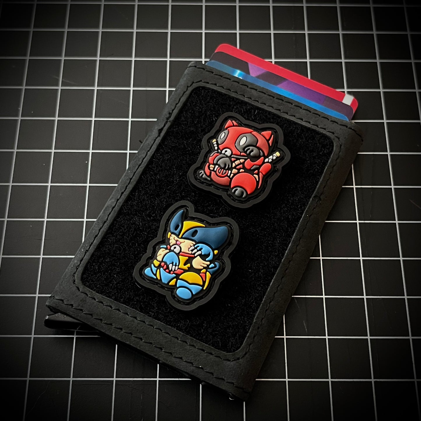 Deadpool & Wolverine Neko RE set PVC ranger eye morale patches of a character in a red suit and another character in a yellow and blue suit sitting in a lucky cat maneki-neko pose on a velcro wallet and a grid background.