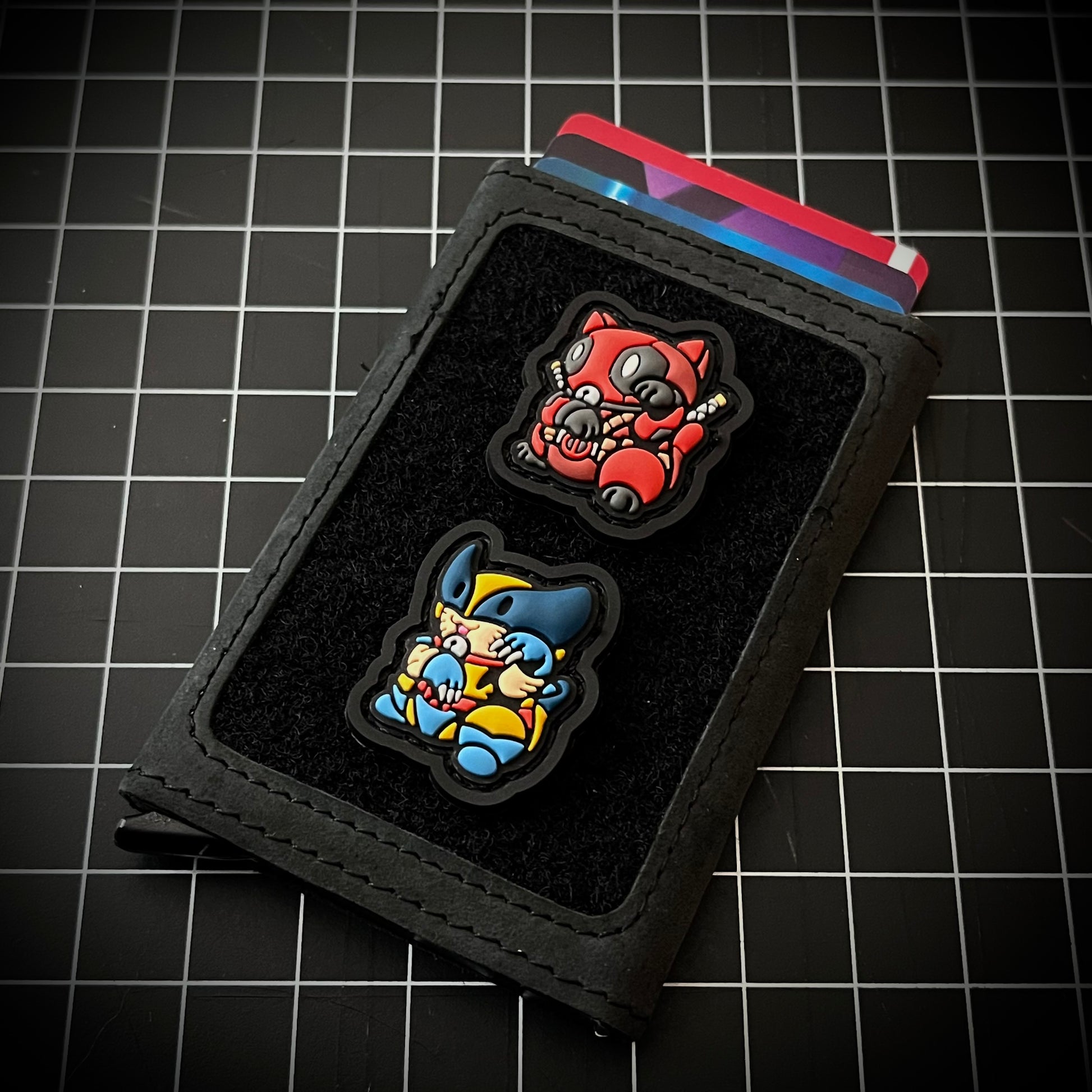Deadpool & Wolverine Neko RE set PVC ranger eye morale patches of a character in a red suit and another character in a yellow and blue suit sitting in a lucky cat maneki-neko pose on a velcro wallet and a grid background.
