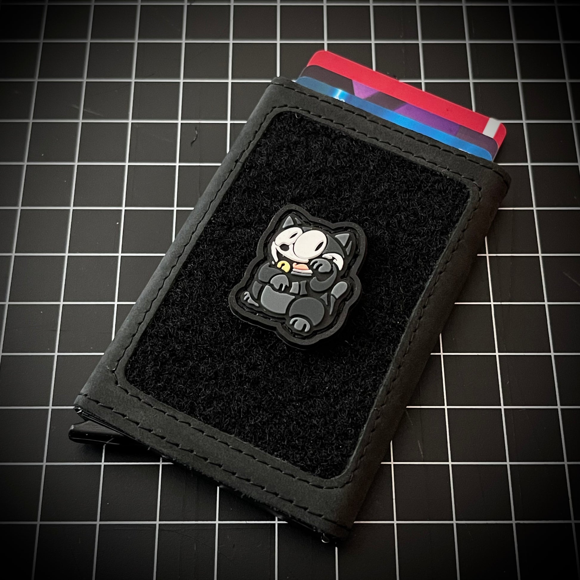 Felix Neko RE PVC ranger eye morale patch of a black cat sitting in a lucky cat maneki-neko pose on a velcro wallet and grid background.