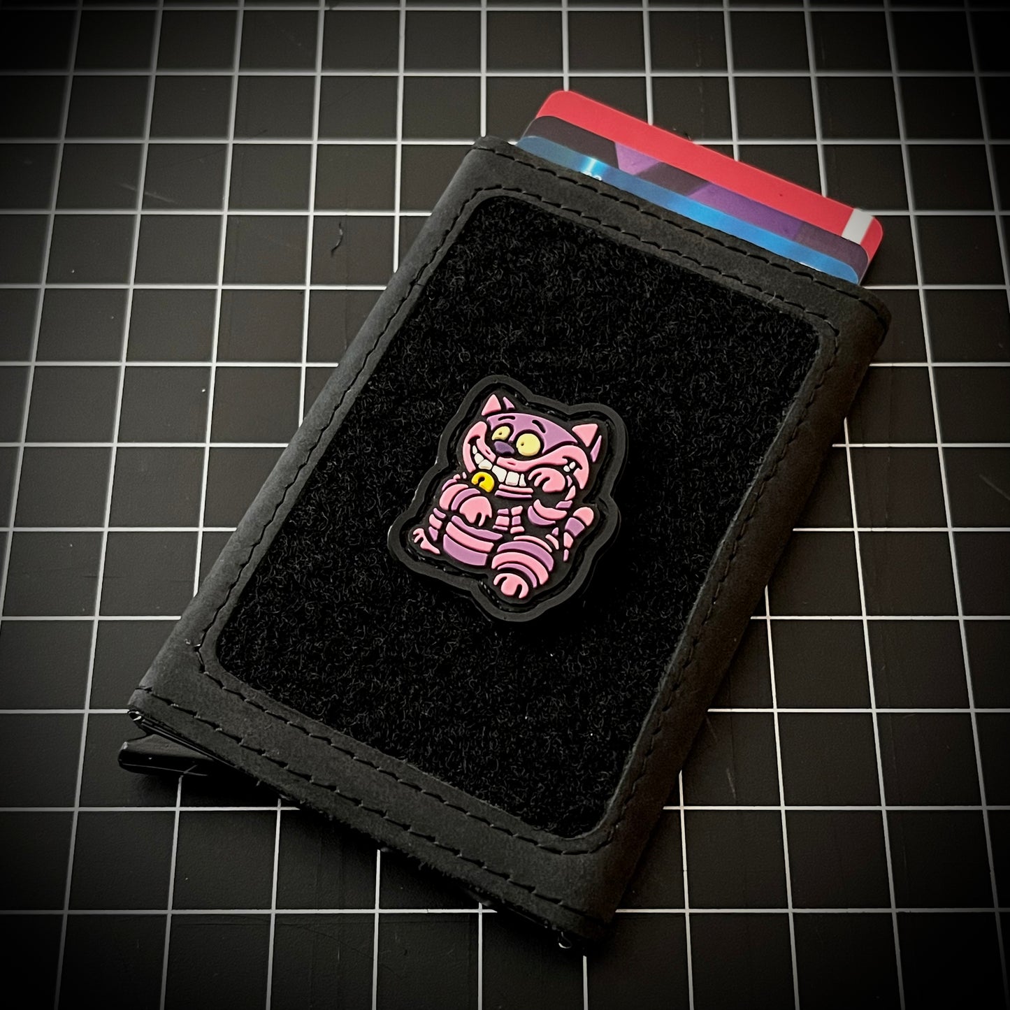 Grinning Cat Neko RE PVC ranger eye morale patch of a purple and pink cat sitting in a lucky cat maneki-neko pose on a velcro wallet and a grid background.