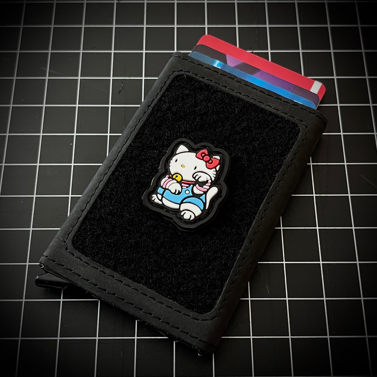 Hello Kitty Neko RE PVC ranger eye morale patch of a white cat wearing a red bowtie and blue clothes sitting in a lucky cat maneki-neko pose on a velcro wallet and grid background.