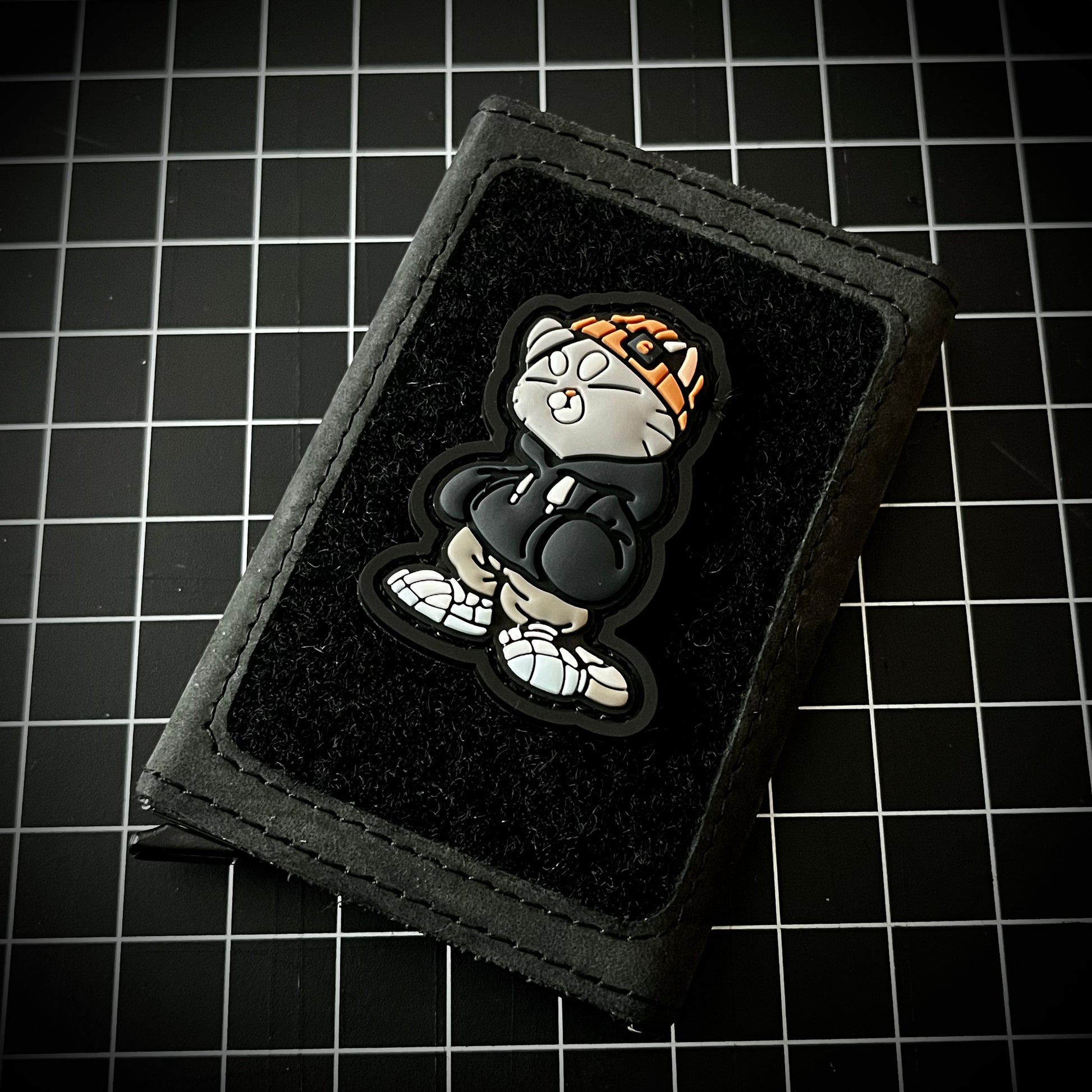 Hoodie Cato Patch PVC ranger eye morale patch of a gray cat in an orange beanie and gray hoodie on a velcro wallet and grid background.