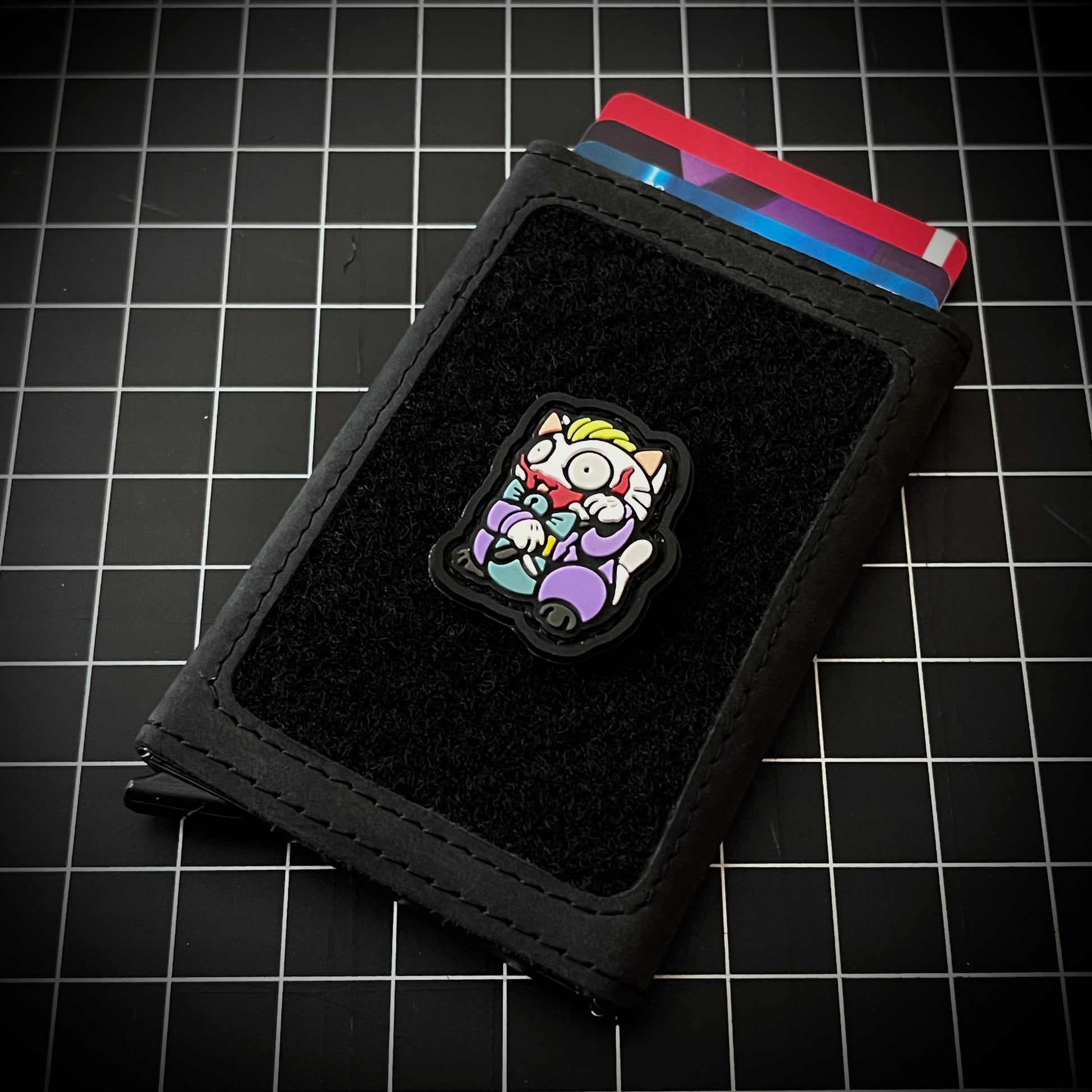 Joker Neko RE PVC ranger eye morale patch of a character wearing a purple suit holding a knife sitting in a lucky cat maneki-neko pose on a velcro wallet and grid background