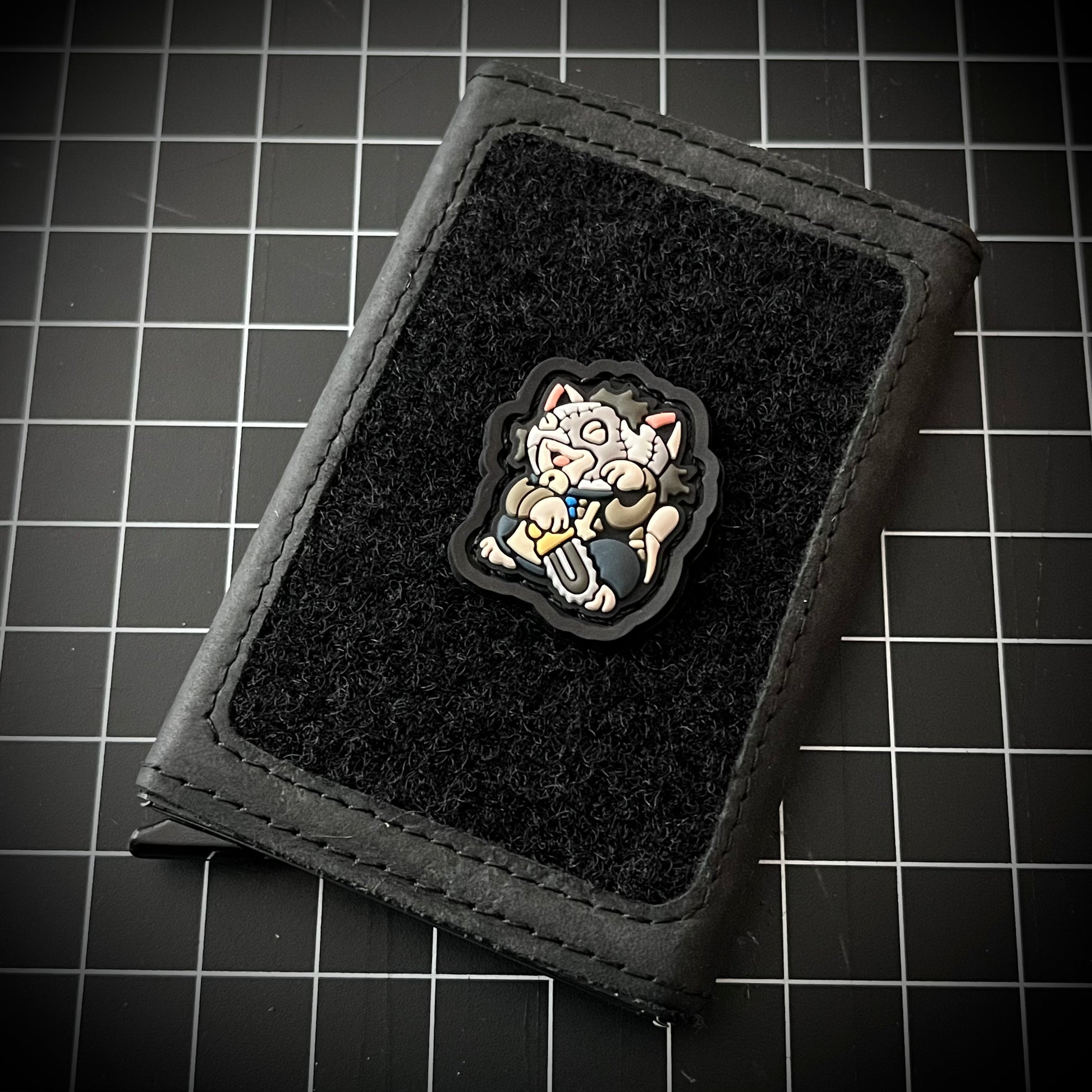 Leatherface Neko RE PVC ranger eye morale patch of a character in a mask holding a chainsaw sitting in a lucky cat maneki-neko pose on a velcro wallet and a grid background.