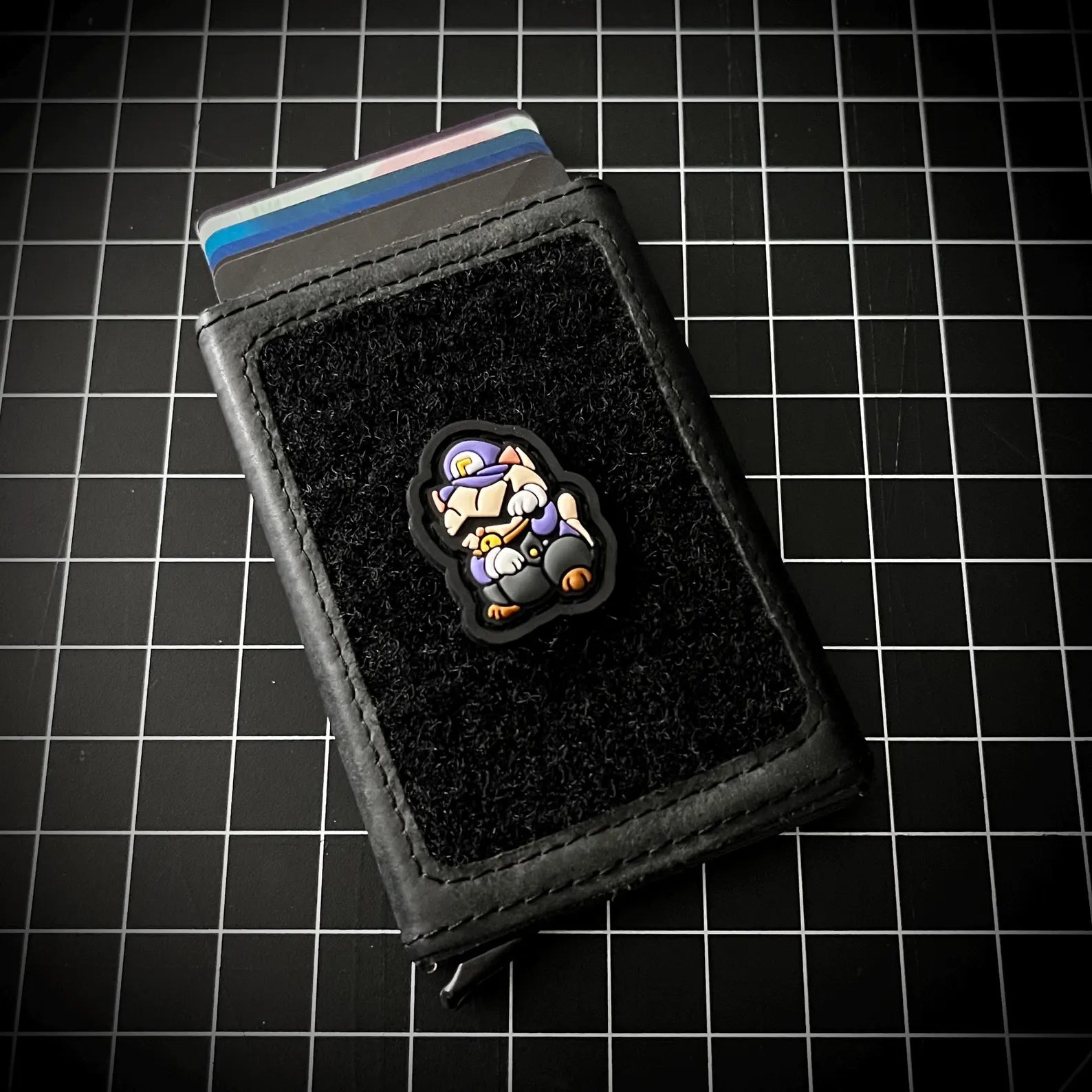 A black leather card holder wallet with a black velcro patch featuring a cartoon plumber character, shown on a black grid background.