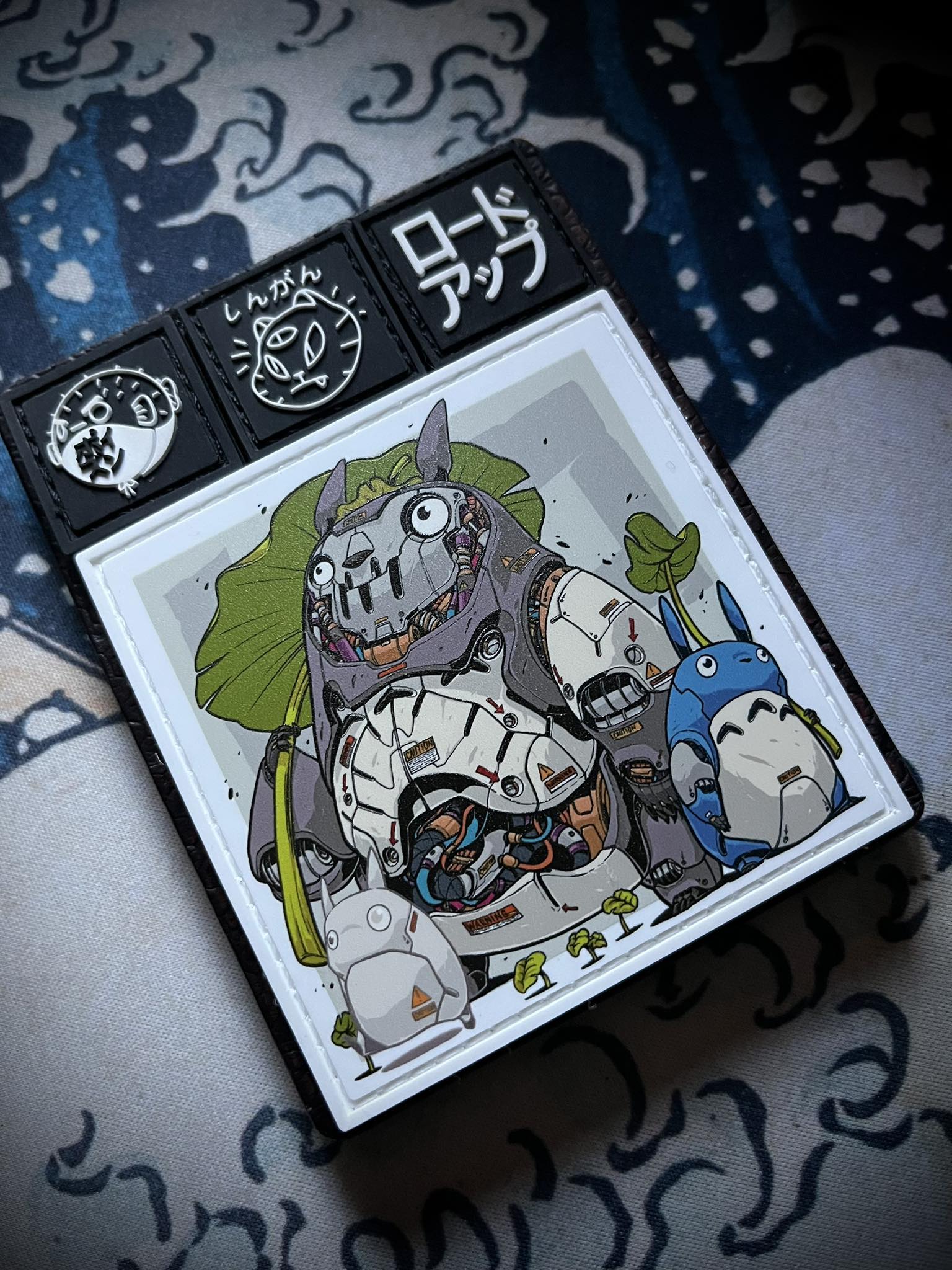 Mechasoul Totoro Patch Cartoon illustration of a large creature with smaller creatures around it on a light gray background and other patches on a wallet on a great wave background