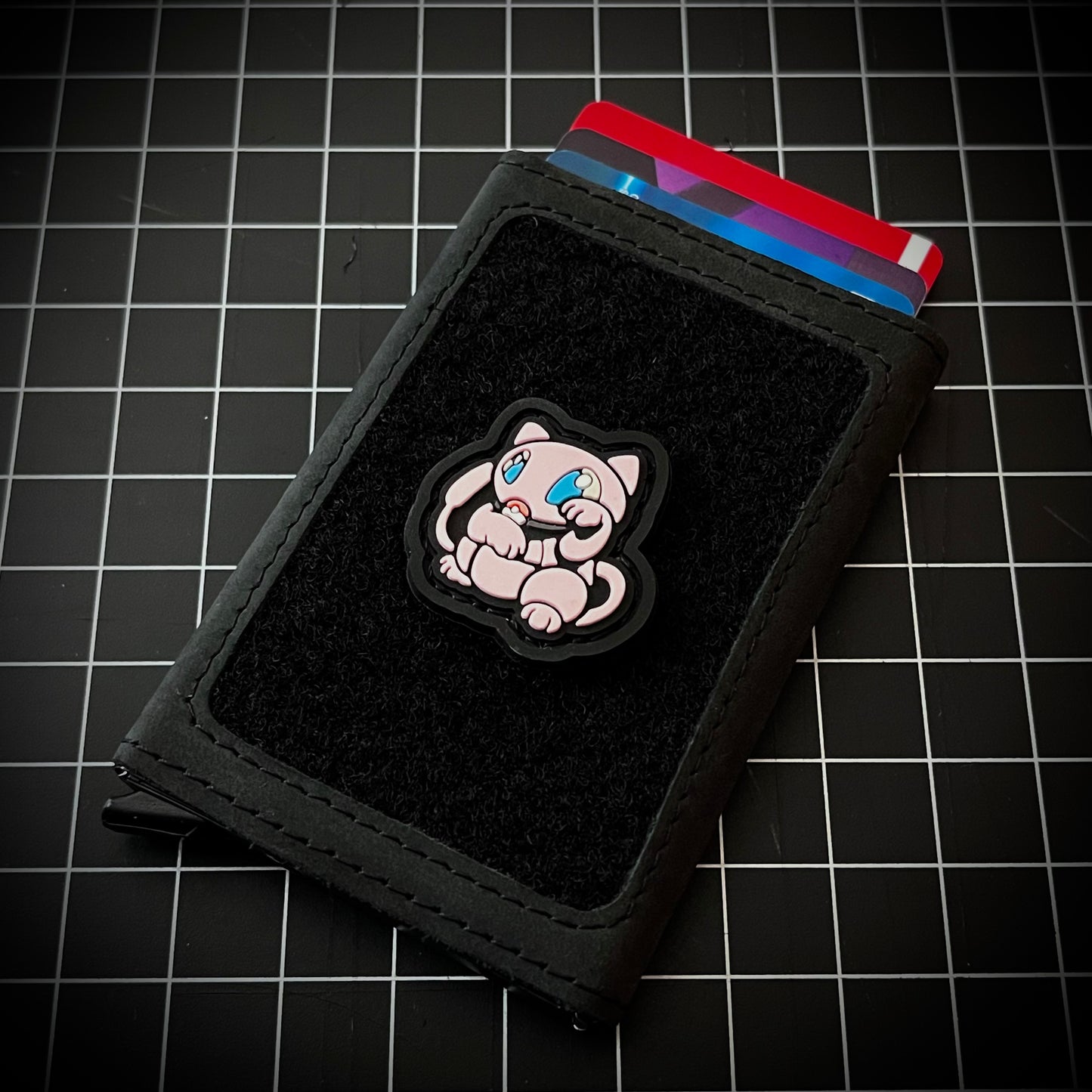Mew Neko RE PVC ranger eye morale patch of a pink cartoon character sitting in a lucky cat maneki-neko pose on a velcro wallet and grid background.