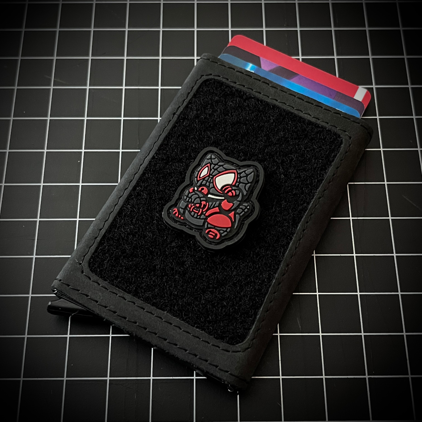 Miles Morales Neko RE PVC ranger eye morale patch of a superhero character in black and red clothing sitting in a lucky cat maneki-neko pose on a velcro wallet and grid background.