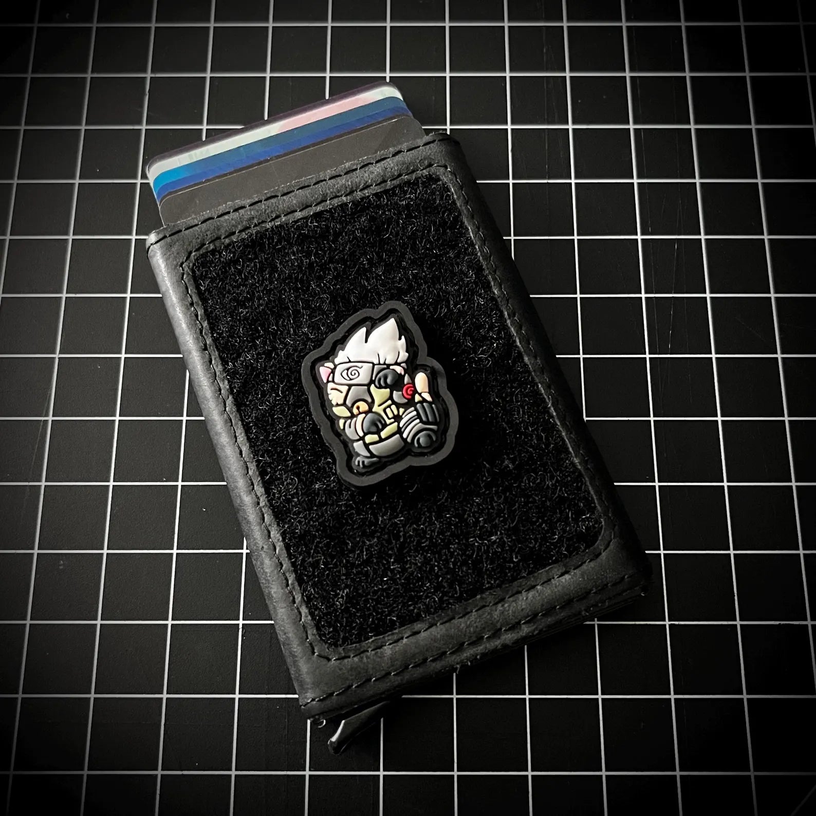 A black leather card holder wallet with a black velcro patch featuring a cartoon character, shown with credit cards inserted on a black grid background.