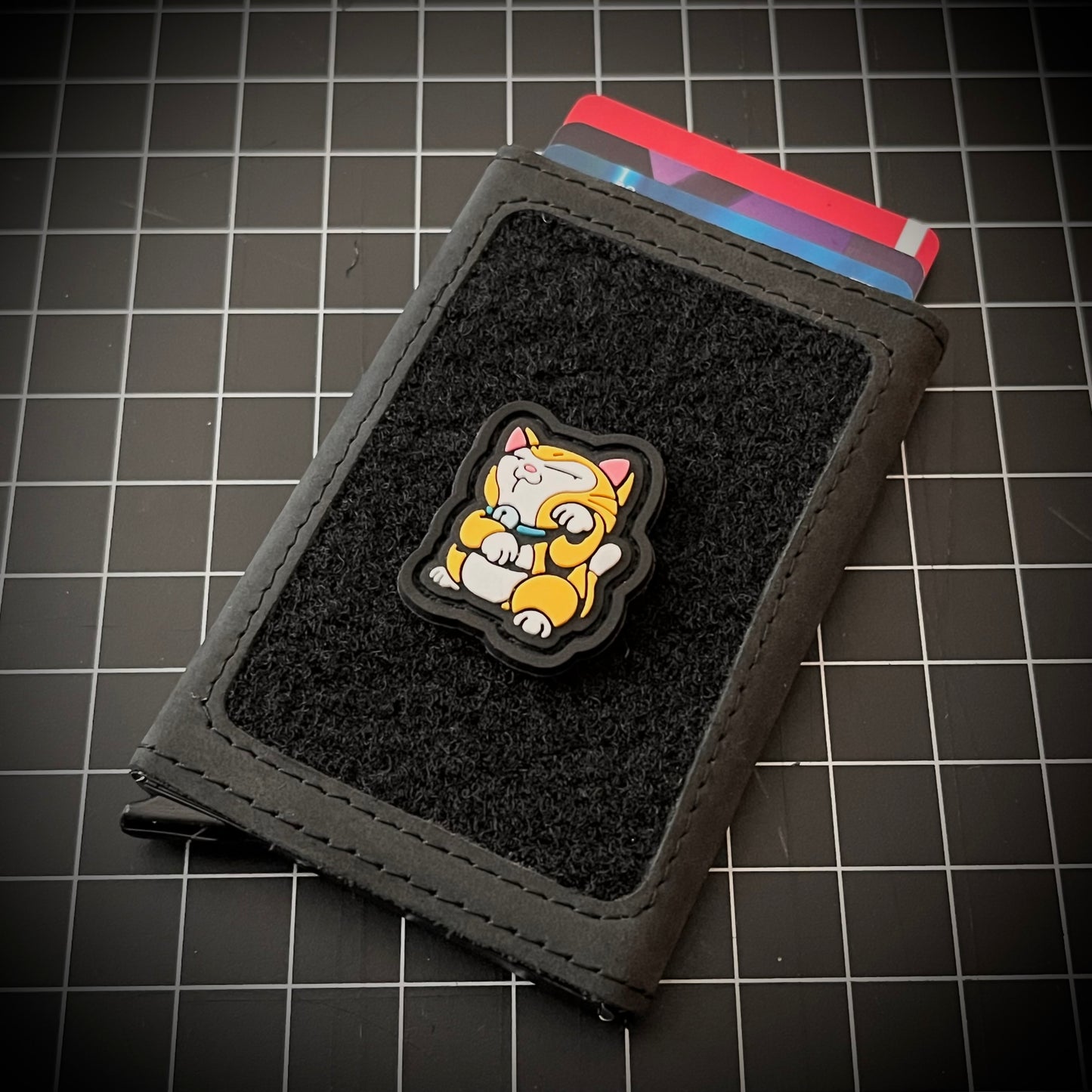 Original Neko RE PVC ranger eye morale patch of an orange cat sitting in a lucky cat maneki-neko on a velcro wallet and grid background.