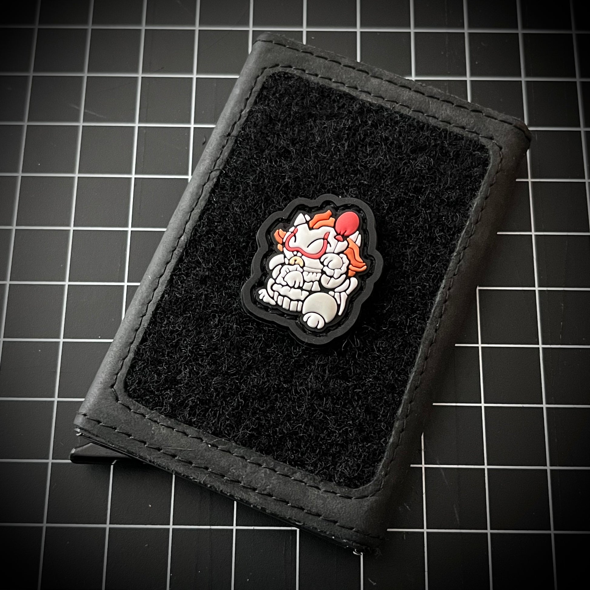 Pennywise Neko RE PVC ranger eye morale patch of a white and orange clown in a lucky cat maneki-neko pose on a velcro wallet and a grid background.