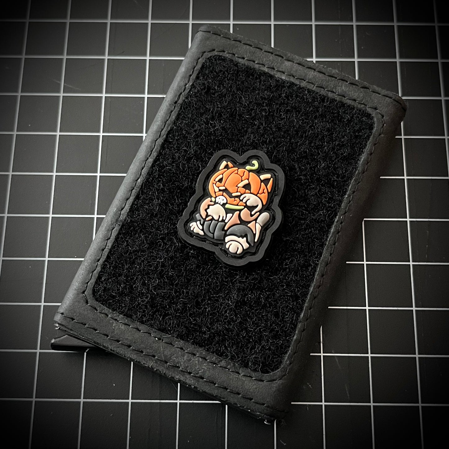 Pumpkin Neko RE PVC ranger eye morale patch of a character with a pumpkin head sitting in a lucky cat maneki-neko pose on a velcro wallet and a grid background.
