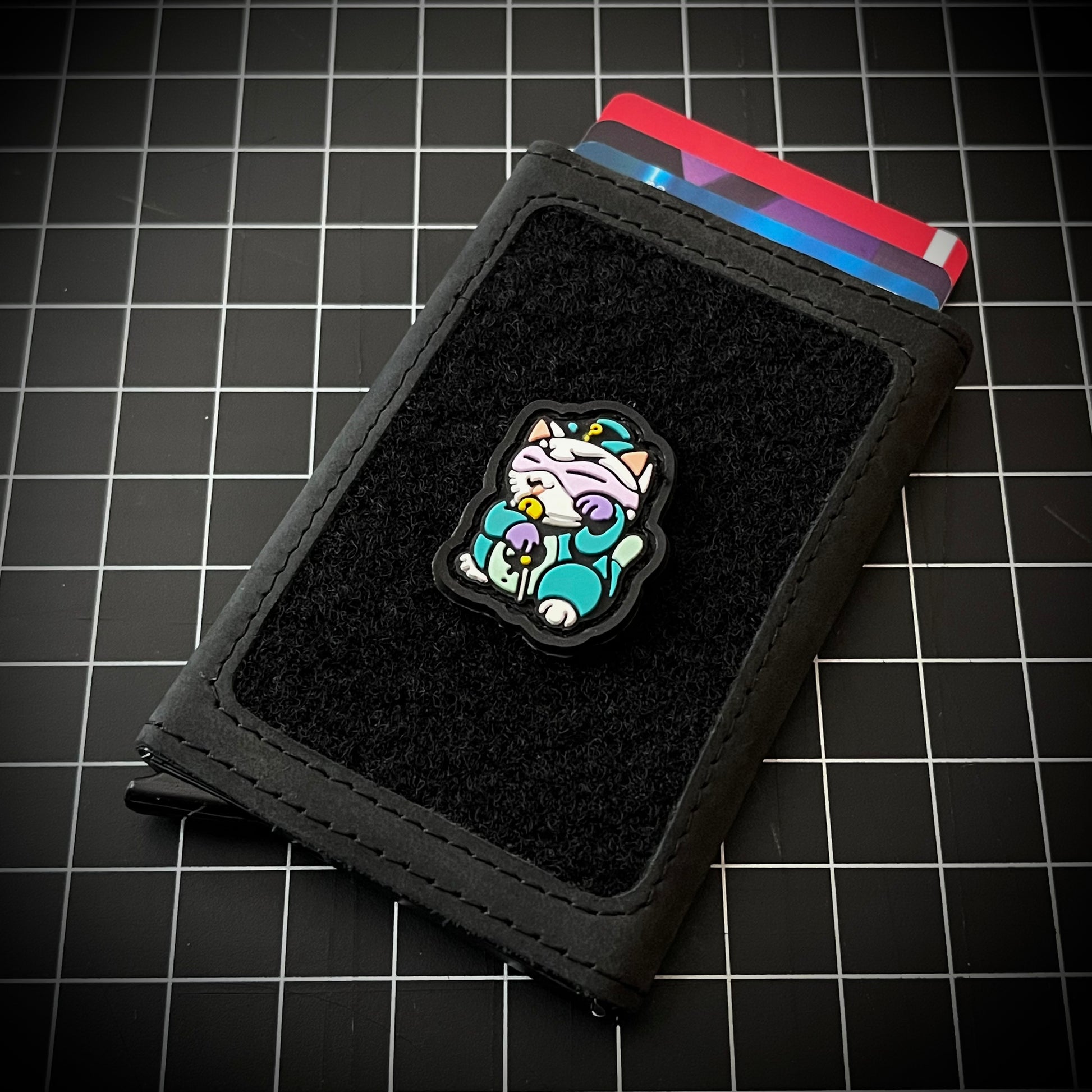 Riddler Neko RE PVC ranger eye neko patch of a character in a purple mask and green outfit sitting in a lucky cat maneki-neko pose on a velcro wallet and grid background.