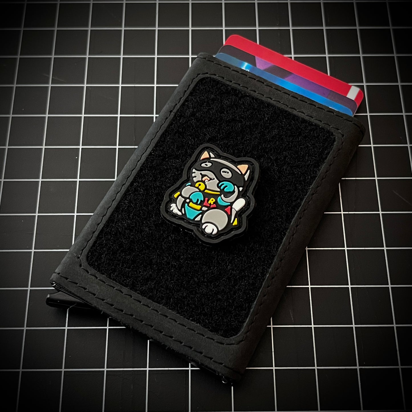 Robin Neko RE PVC ranger eye morale patch of a gray cat wearing a mask and sidekick outfit in a lucky cat maneki-neko pose on a velcro wallet and a grid background.