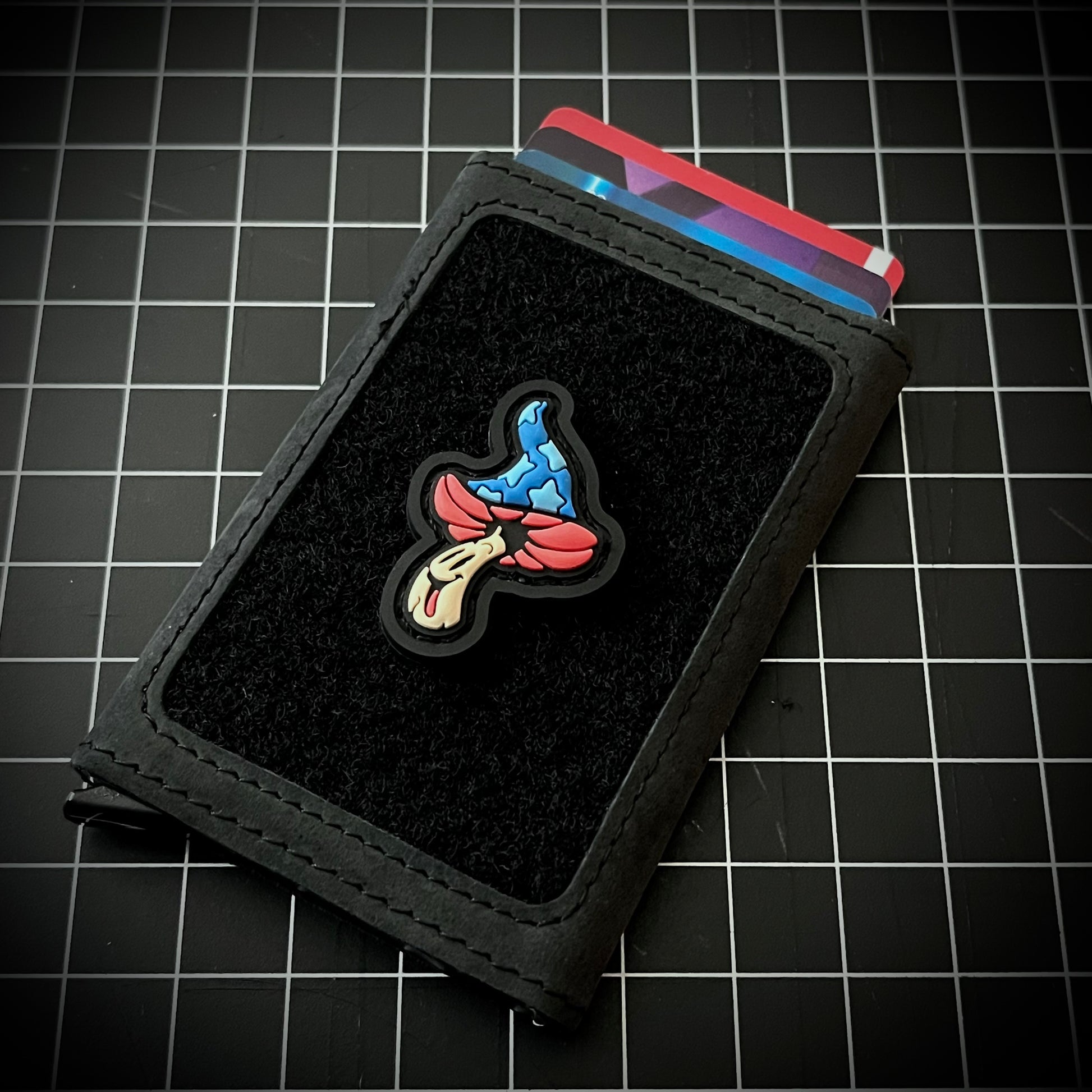 Shroomie RE - Fantasia PVC ranger eye morale patch of a red, blue, and white smiling mushroom whose cap is reminiscent of mickey sorcerer fantasia on a velcro wallet and a grid background