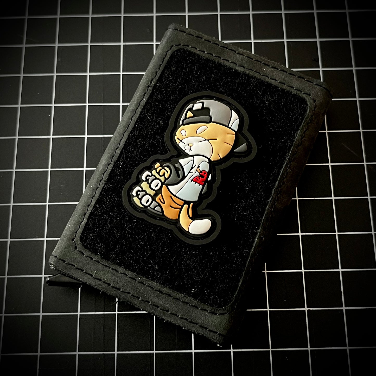 Skate Cato RE PVC ranger eye morale patch of an orange cat wearing a backwards hat and Toy Machine shirt holding a skateboard on a velcro wallet and a grid background.