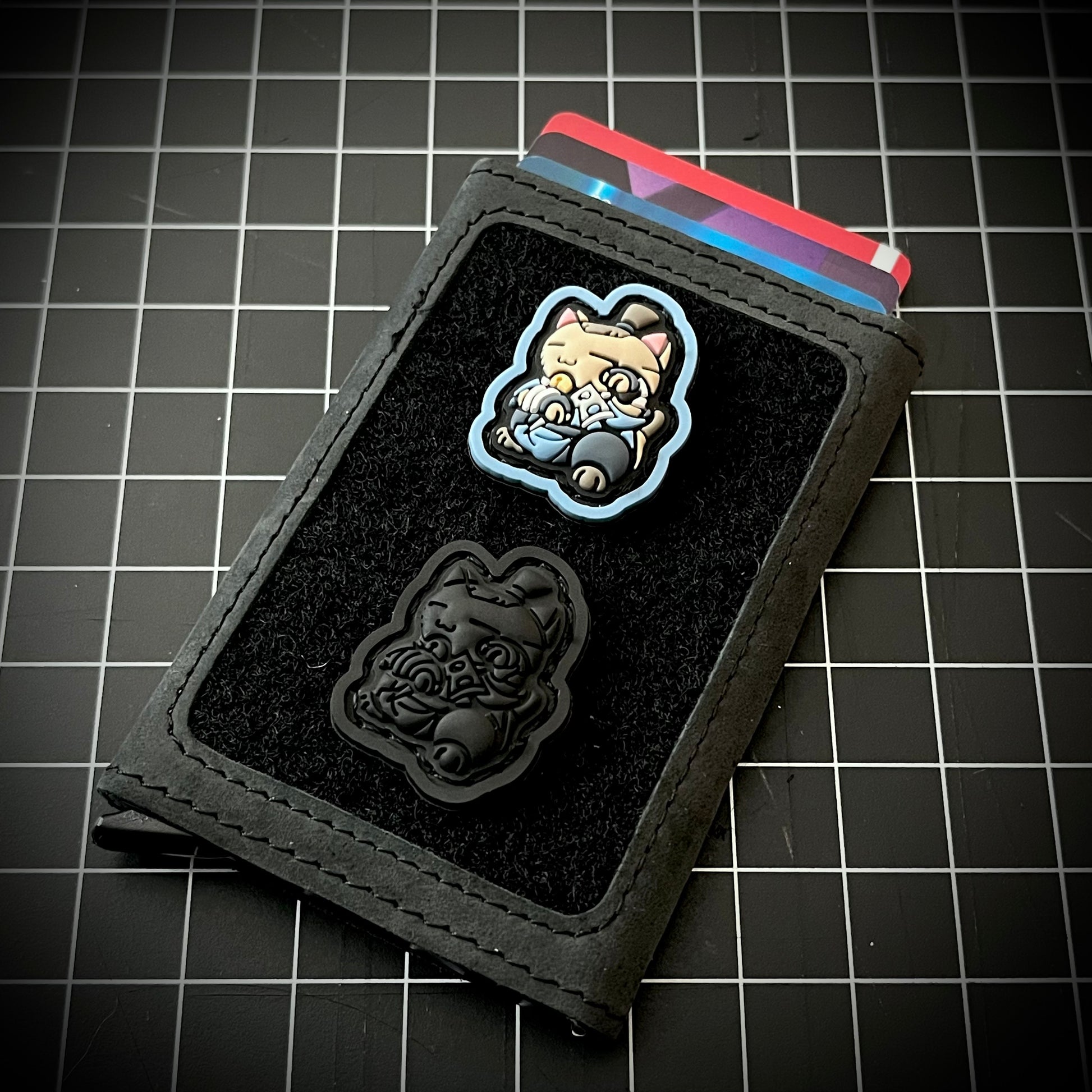 Sokka Neko RE and Sokka Neko Blackout RE PVC ranger eye patches of a cartoon character on a velcro wallet with a grid background.