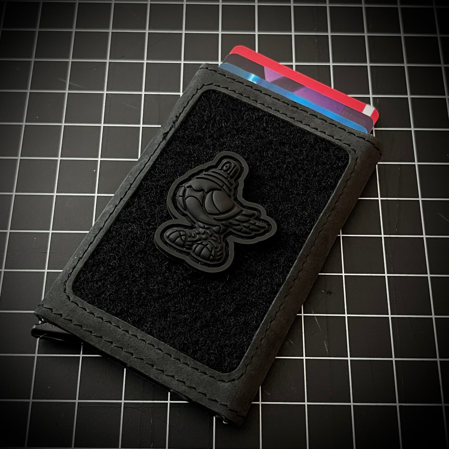 Spraygeon RE - Blackout PVC ranger eye morale patch of a fully black pigeo with spray paint can tip for a head and wearing tennis shoes on a velcro wallet and grid background.