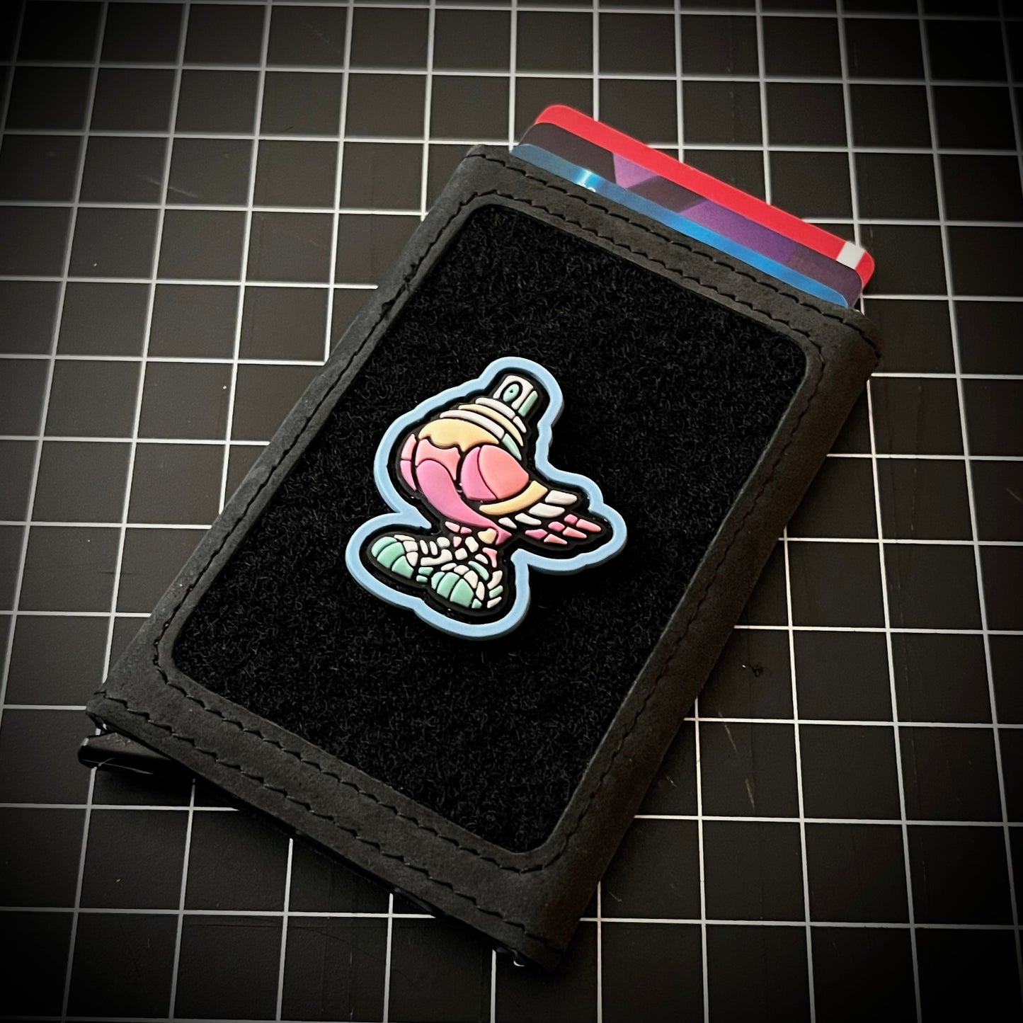 Spraygeon RE - Candy PVC ranger eye morale patch of a pink pigeon with a spray paint cap head and tennis shoes with a blue outline on a velcro wallet and grid background.