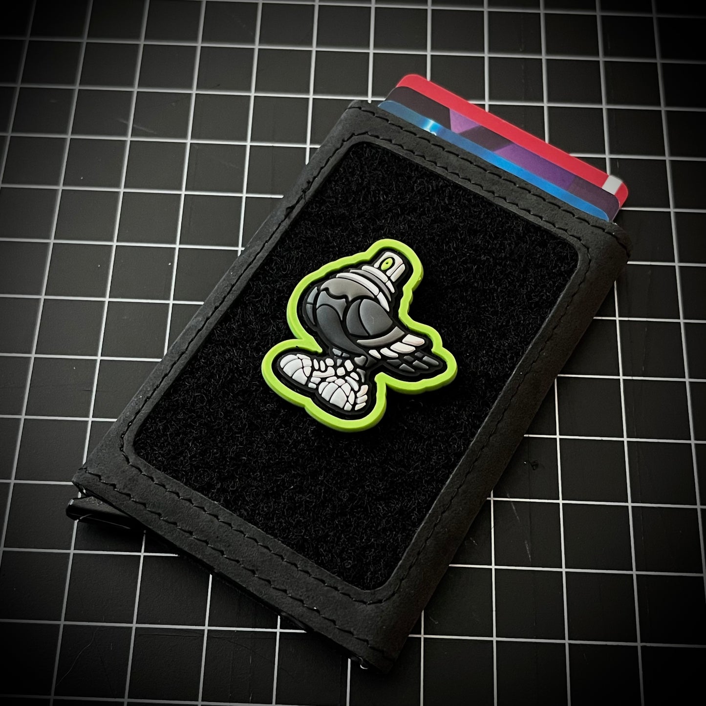 Spraygeon RE PVC ranger eye morale patch of a gray pigeon character with a spray can cap head and tennis shoes and lime green outline on a velcro wallet and grid background.