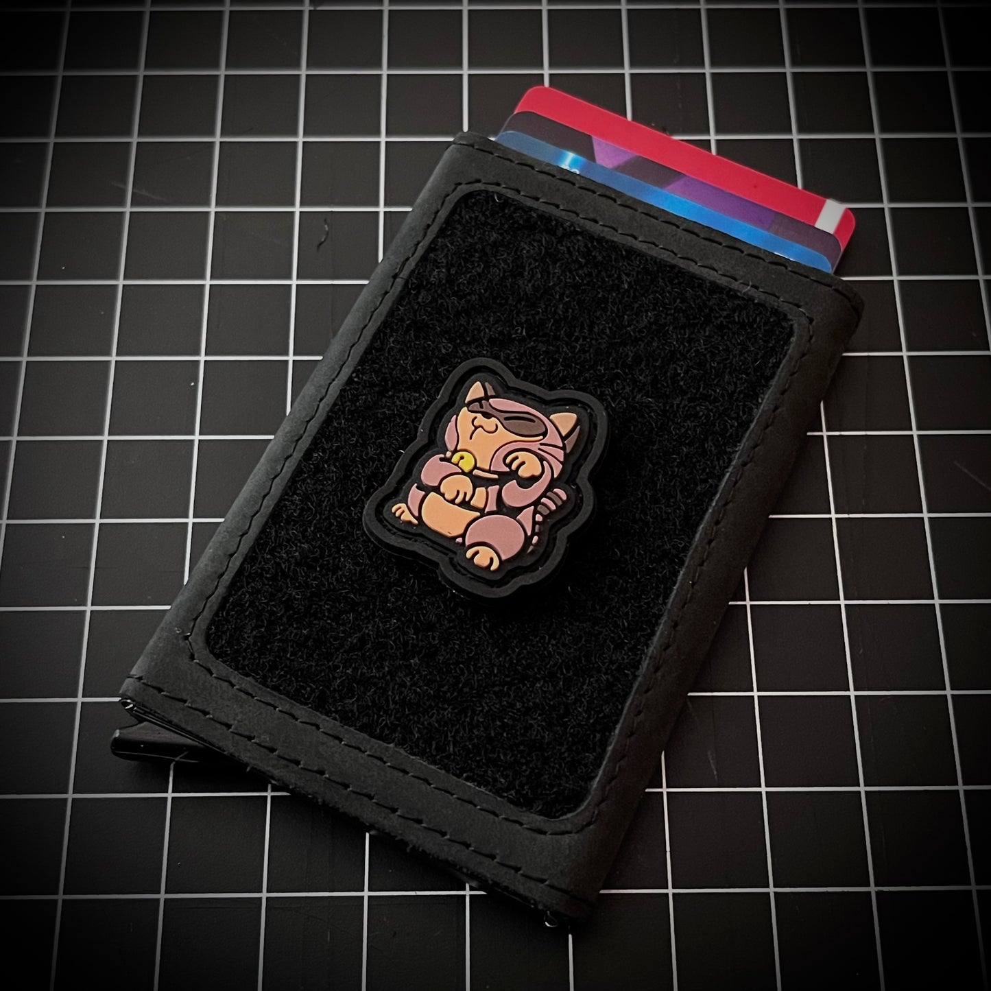 Tanooki Neko RE PVC ranger eye patch of a brown game character in a lucky cat maneki-neko pose on a velcro wallet and grid background.