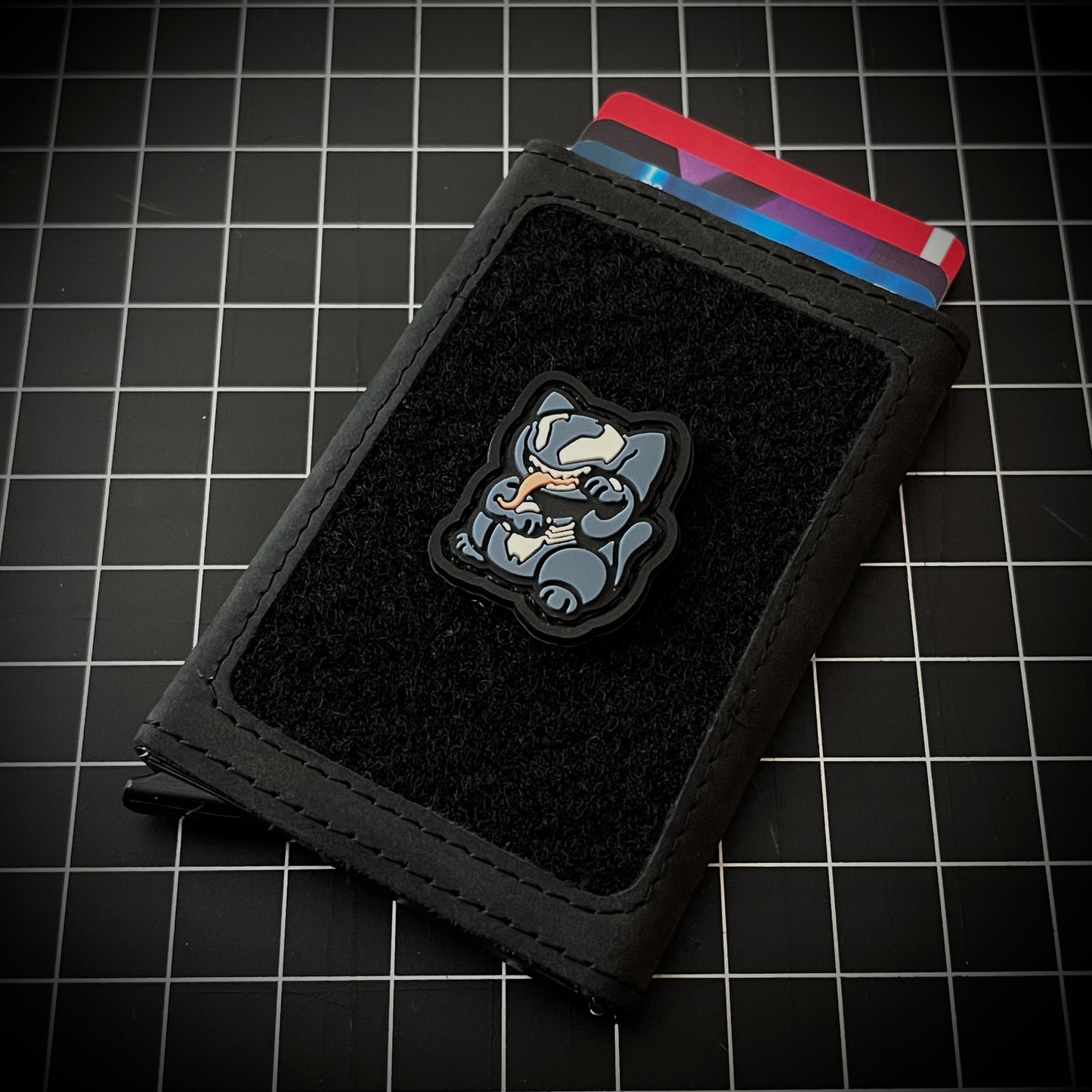 Venom Neko RE PVC ranger eye morale patch of a blue cartoon character with its tongue sticking out sitting in a lucky cat maneki-neko pose on a velcro wallet and grid background.